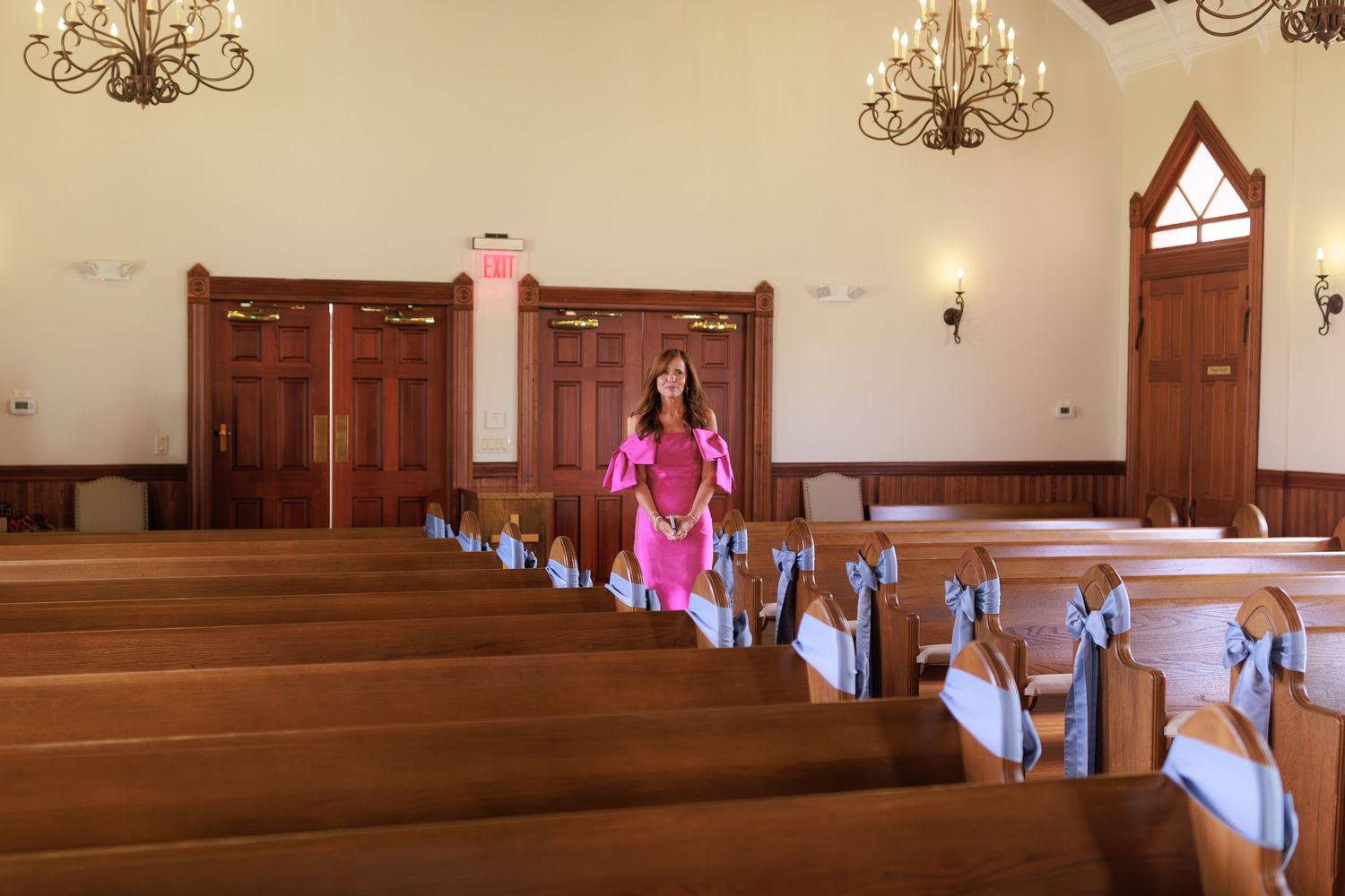 Person in bright pink dress walking down church aisle decorated with powder blue bows on wooden pews.