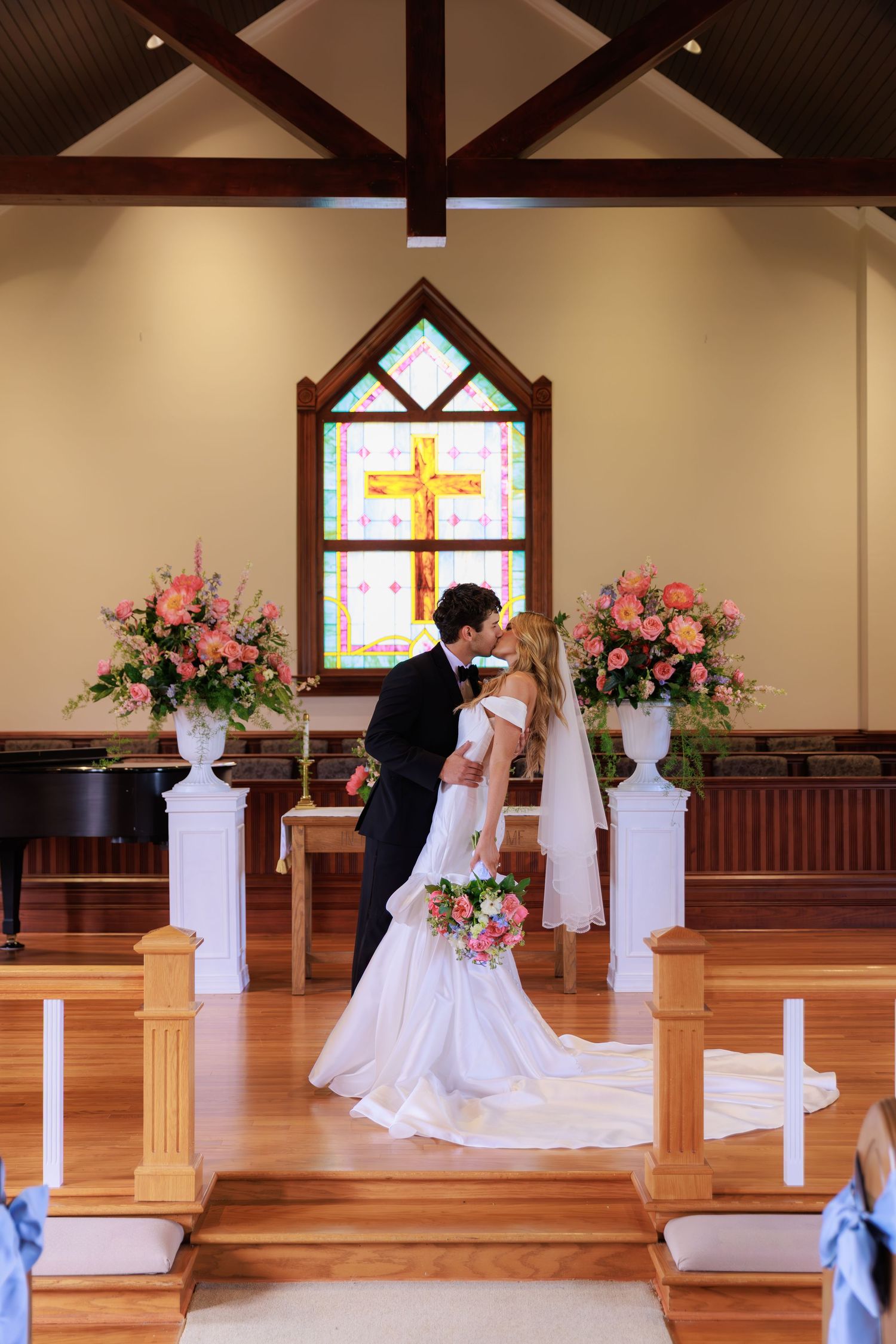 Elegant wedding ceremony in a chapel with stained glass cross window and pink floral arrangements on white pedestals.