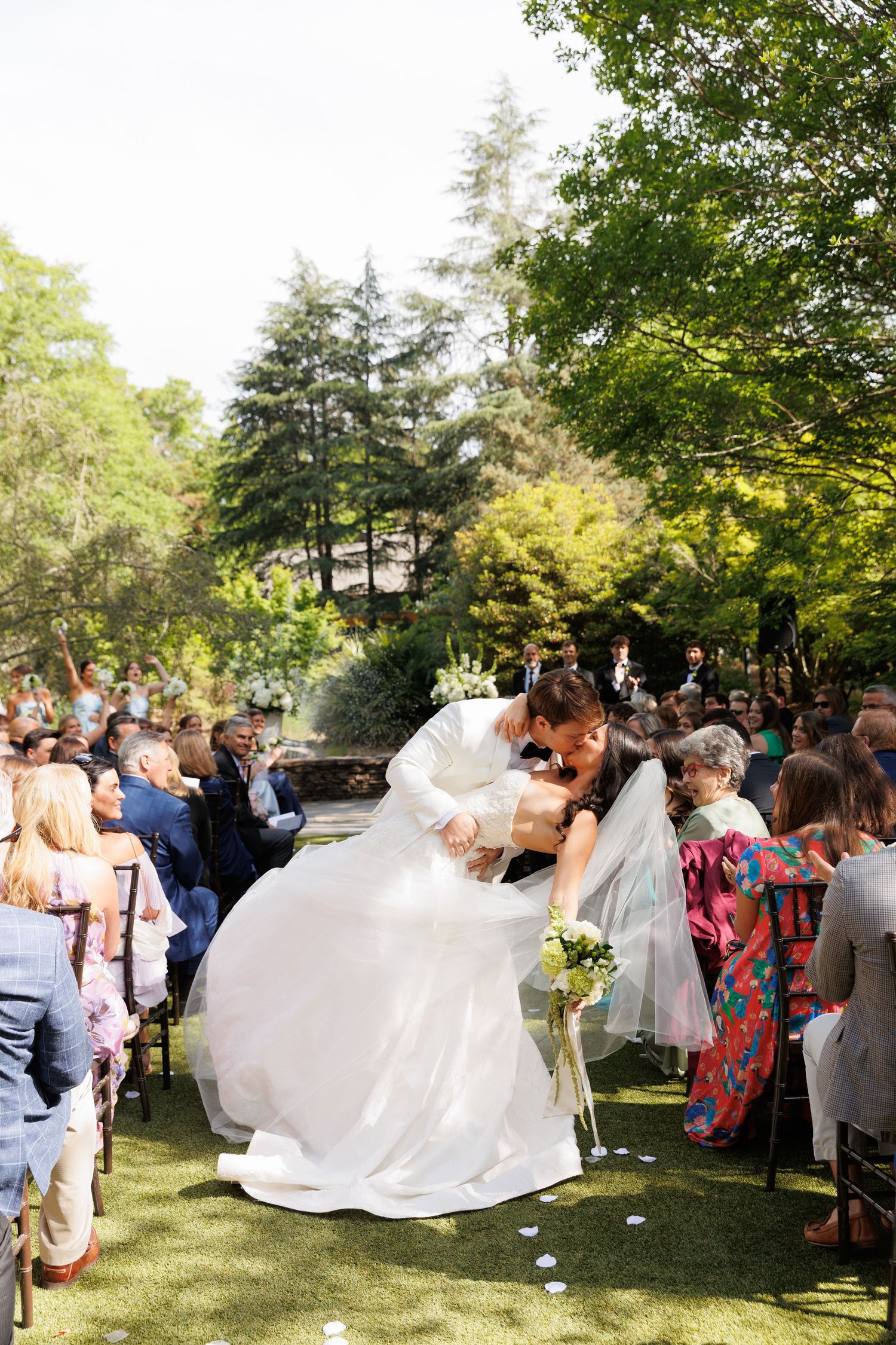 Groom dips bride in white wedding dress for a kiss along outdoor garden aisle lined with guests.