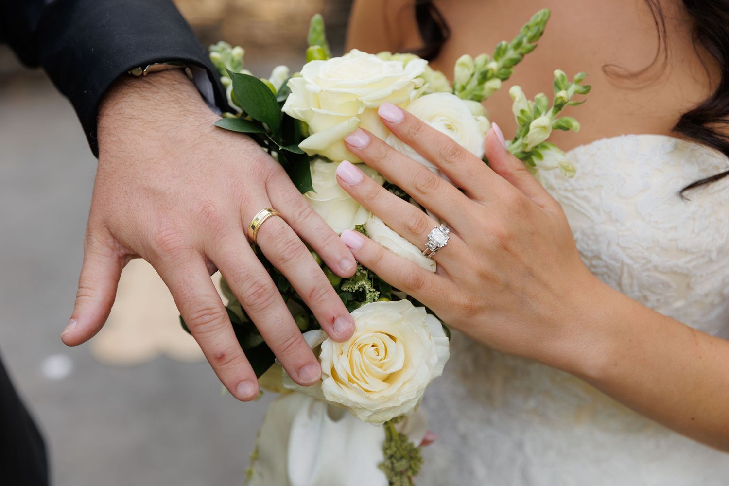 Bride and groom displaying wedding rings on hands resting over a bouquet of white roses and greenery.