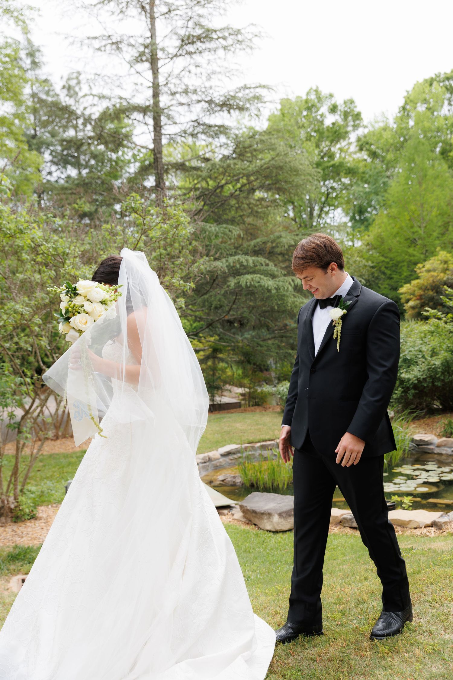Groom in black tuxedo looks down at bride in white dress with bouquet in garden.