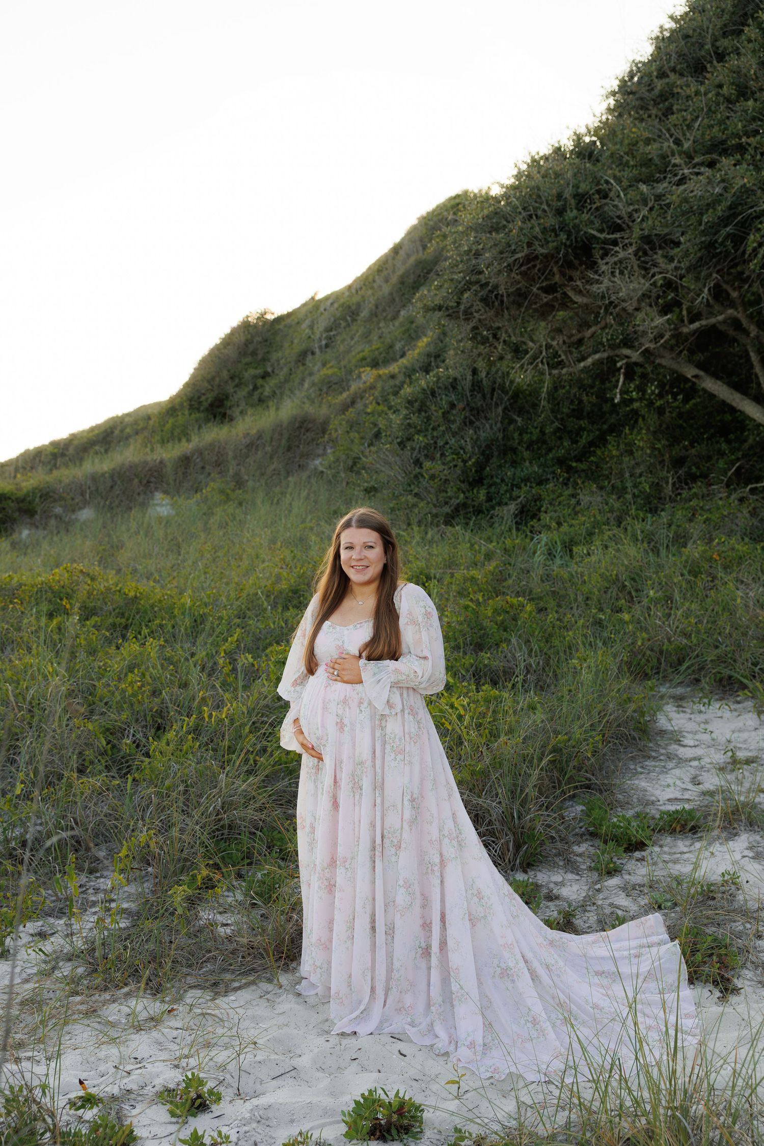 Pregnant woman in flowing floral gown poses on sandy beach dunes surrounded by lush green vegetation at golden hour.