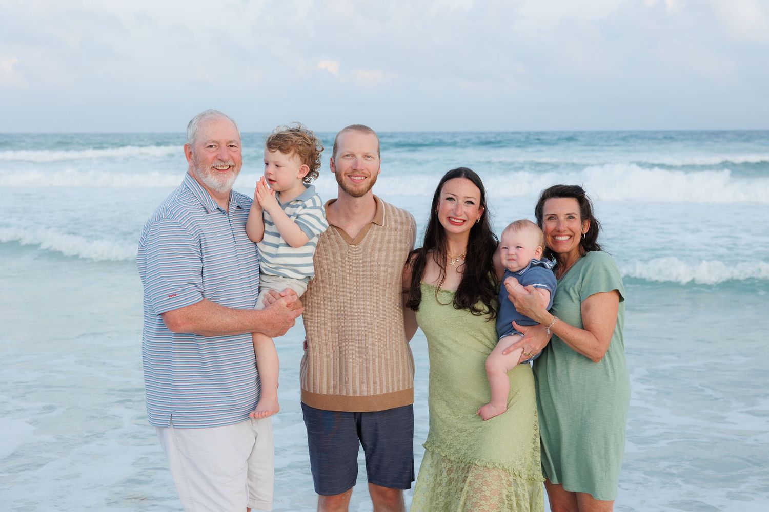 Multi-generational family of six smiling together on a sandy beach with ocean waves in the background.