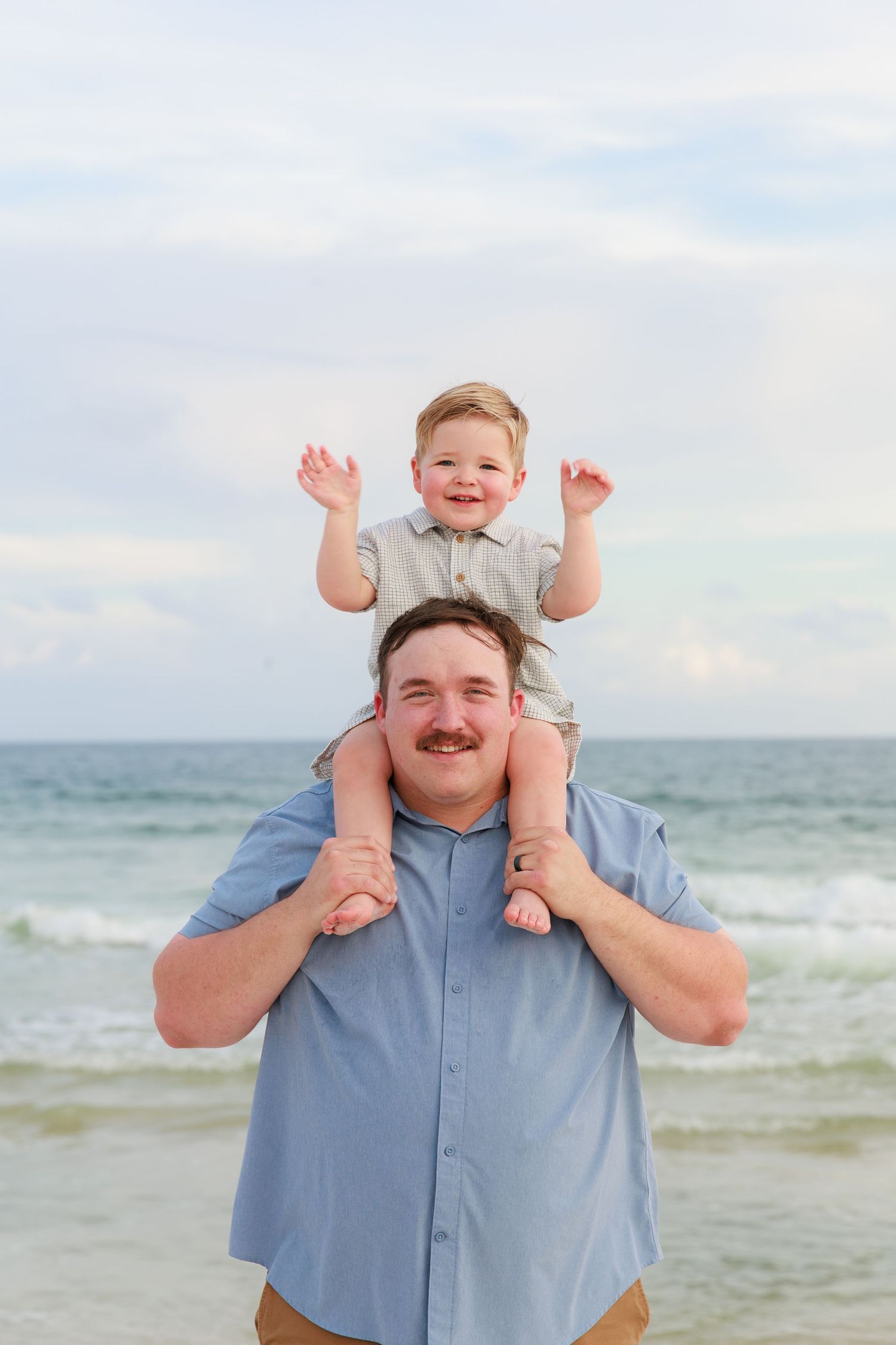 A joyful moment shared between parent and child playing on shoulders at the beach with ocean waves in background.