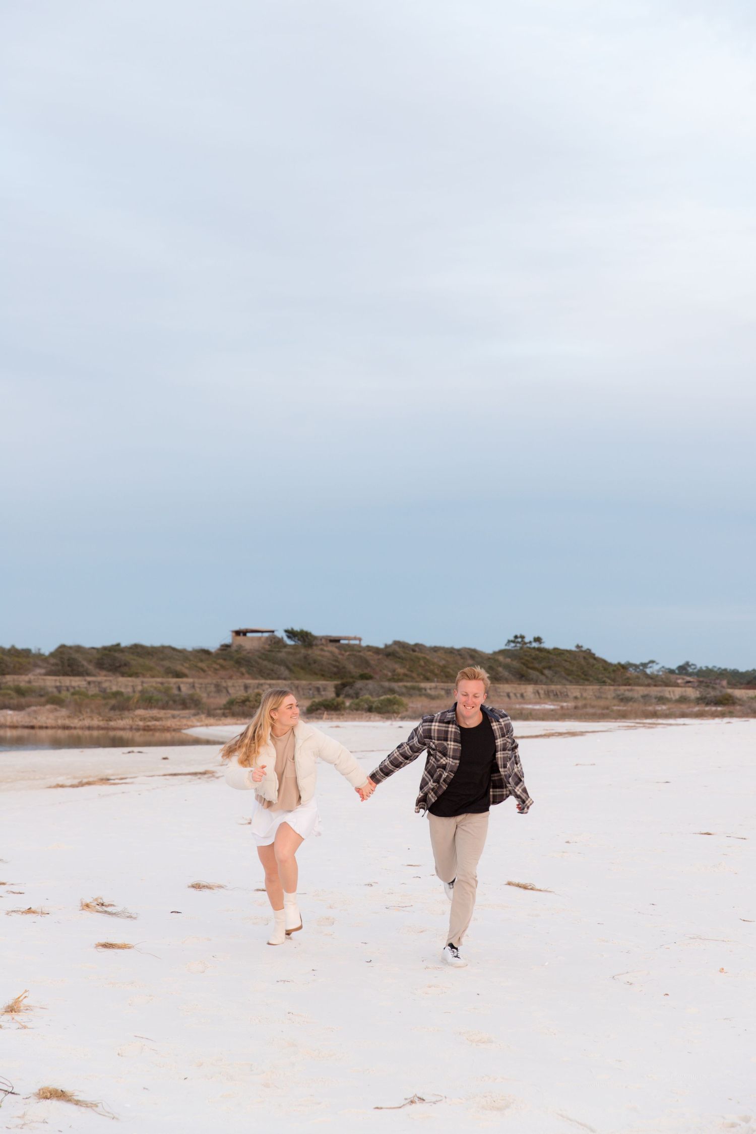 A couple walks hand in hand along a pristine white sandy beach with dunes in the background on a cloudy day.