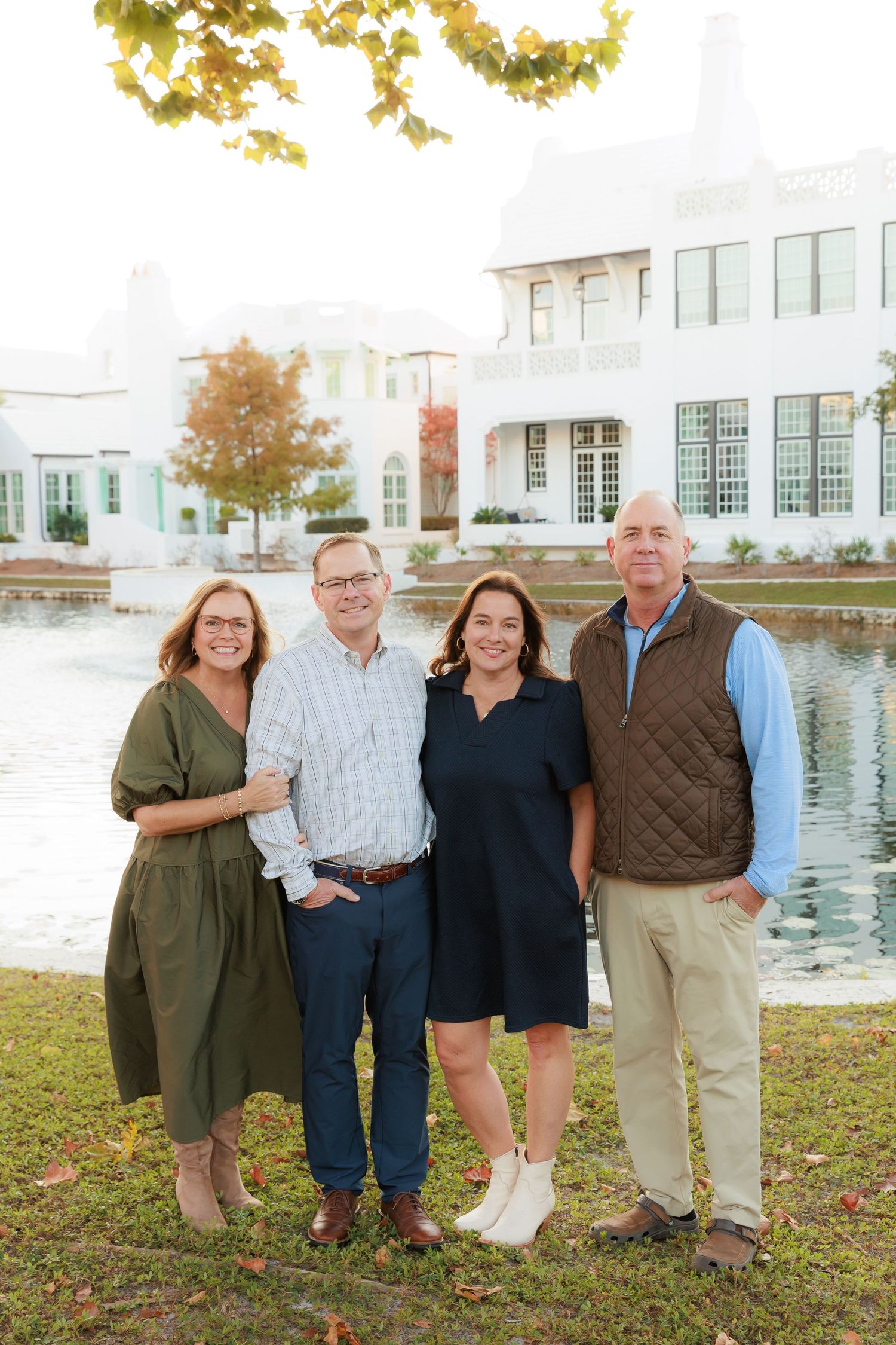 A group stands together on a grassy lawn near a waterfront with autumn trees and white buildings in the background.