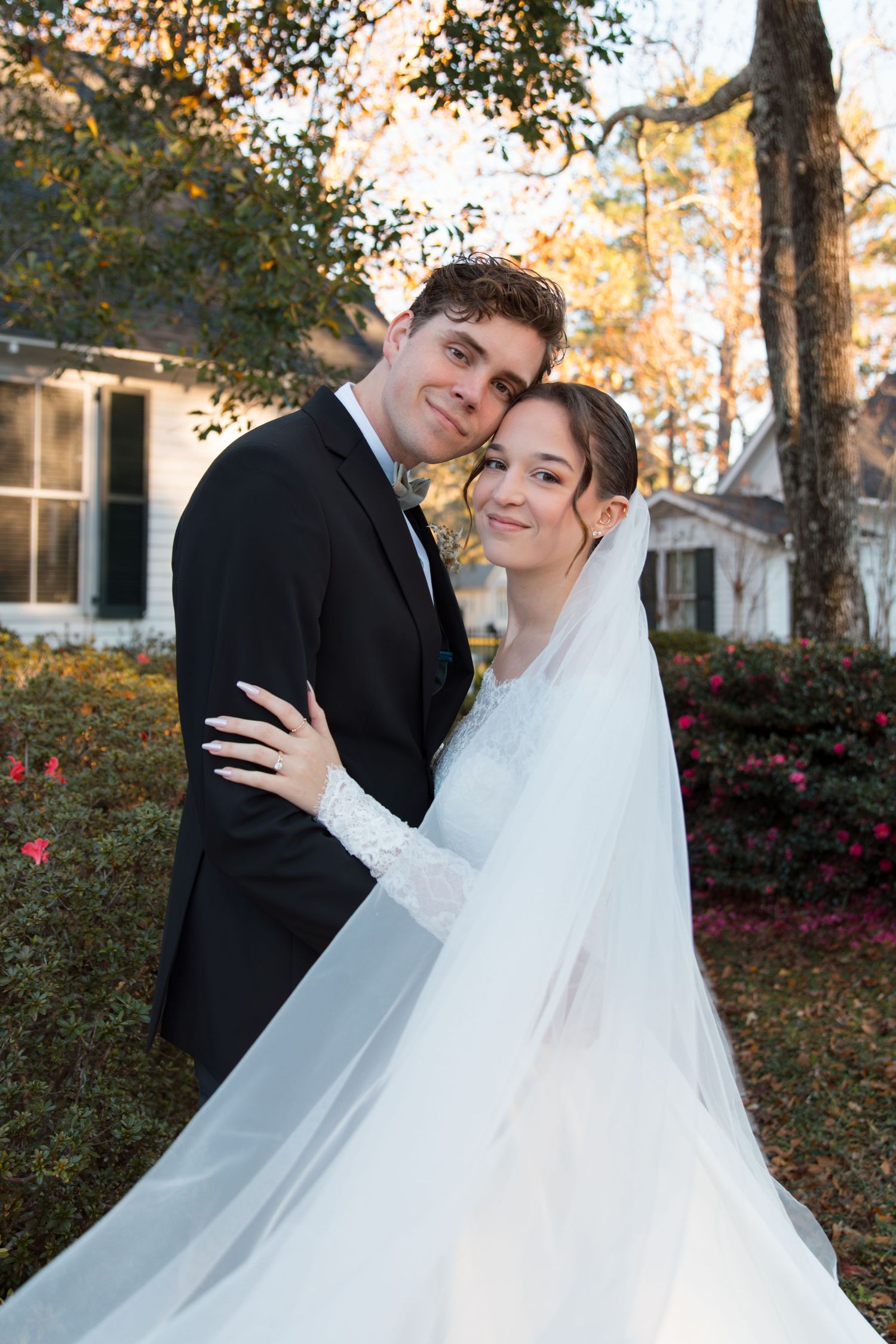 A bride in a flowing white dress and veil embraces her groom in a black suit during an autumn outdoor wedding photoshoot.