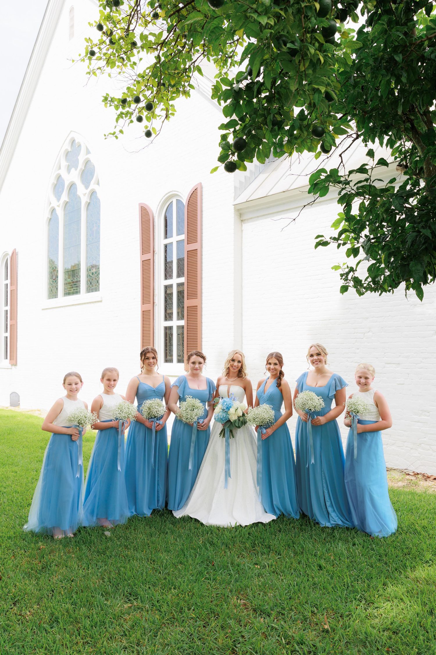 Large bridal party in blue dresses poses with bride in white gown outside historic church.