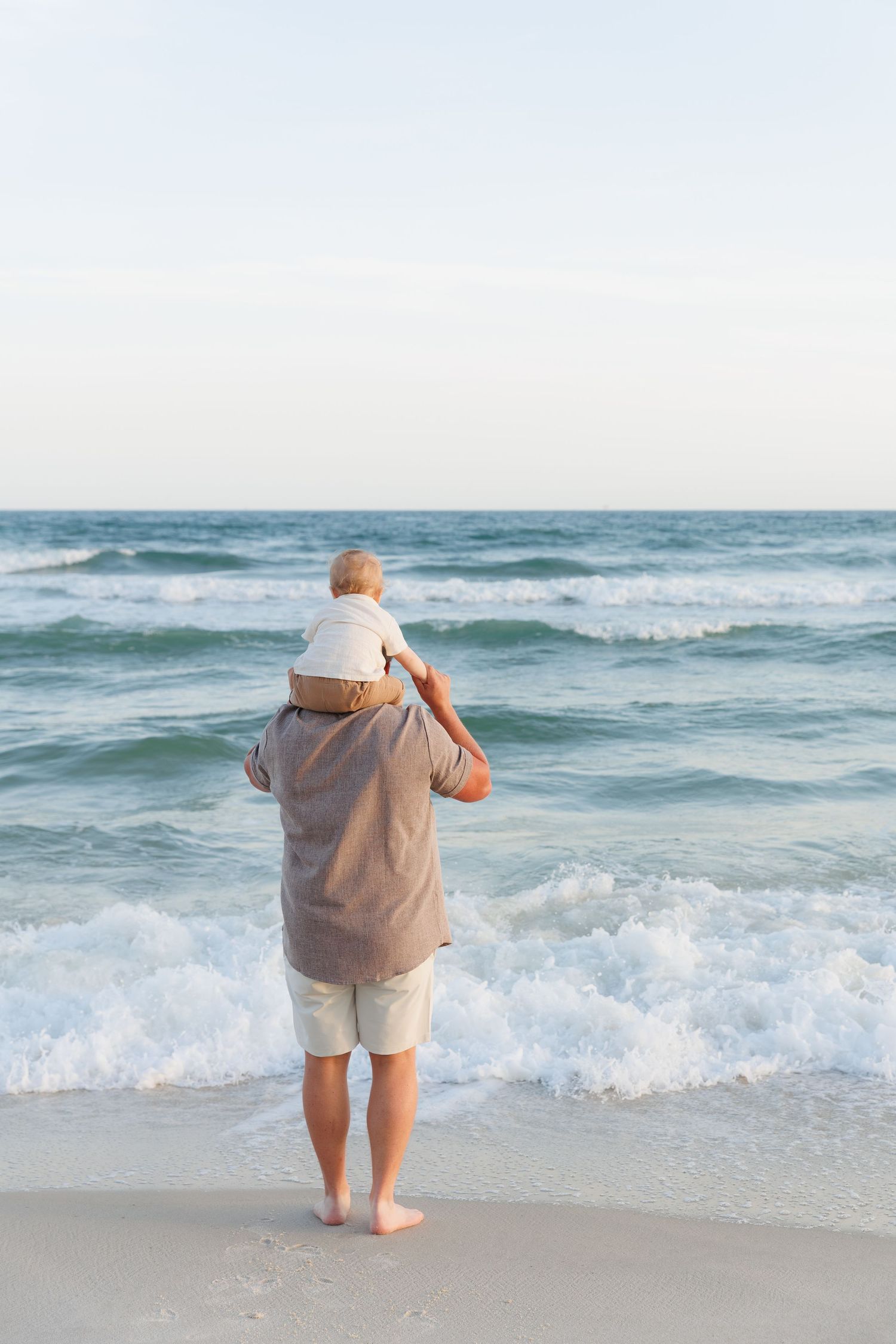 Man carrying baby on shoulders standing barefoot at ocean's edge on a sunny beach.