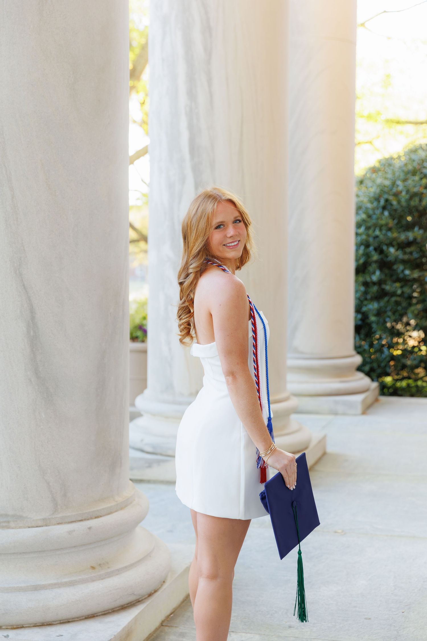 Female graduate in white dress holds cap with green tassel, wearing honor sash between elegant columns.