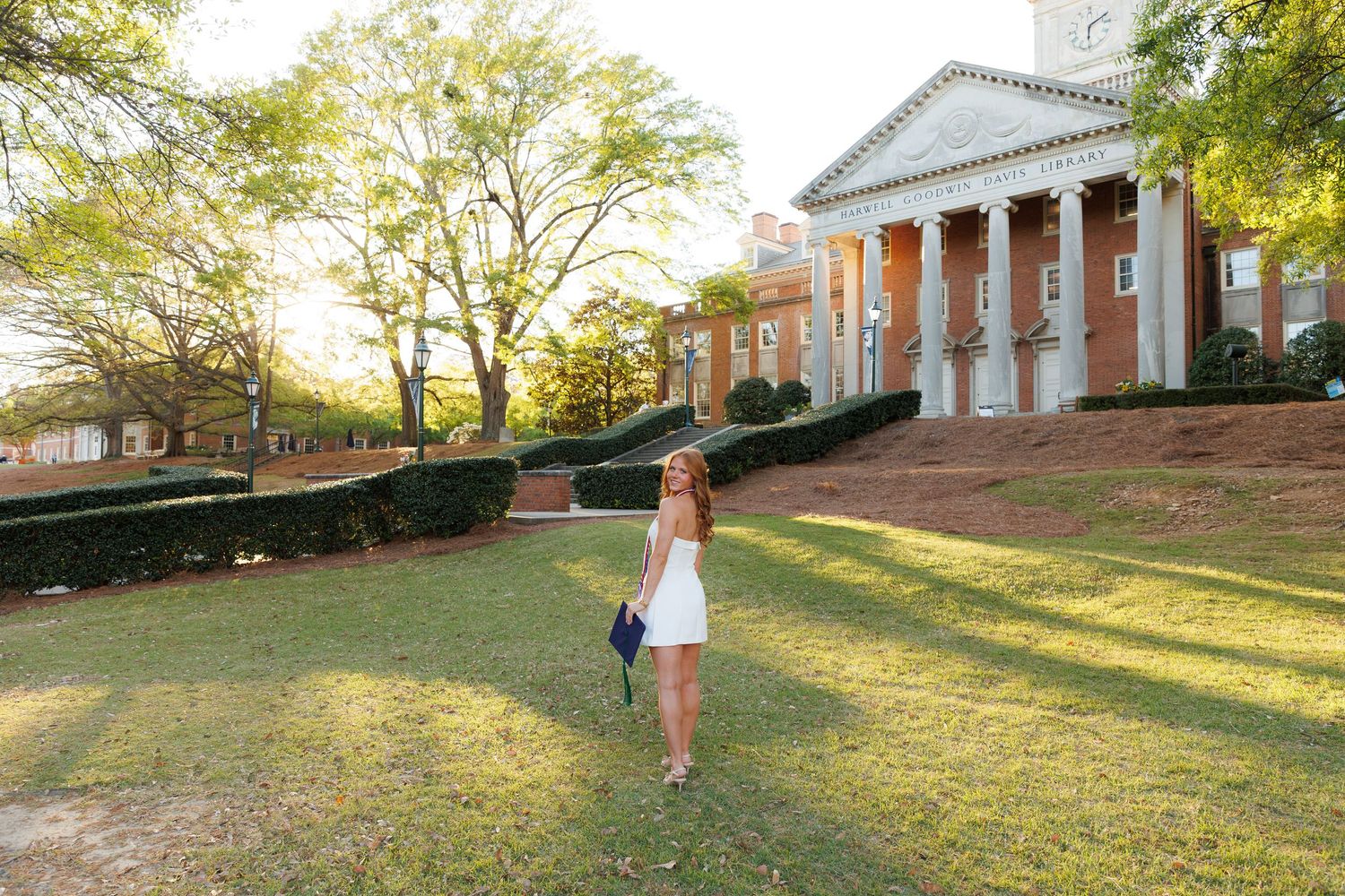 Young woman in white dress walking on green lawn in front of a classic brick colonial building with white columns.