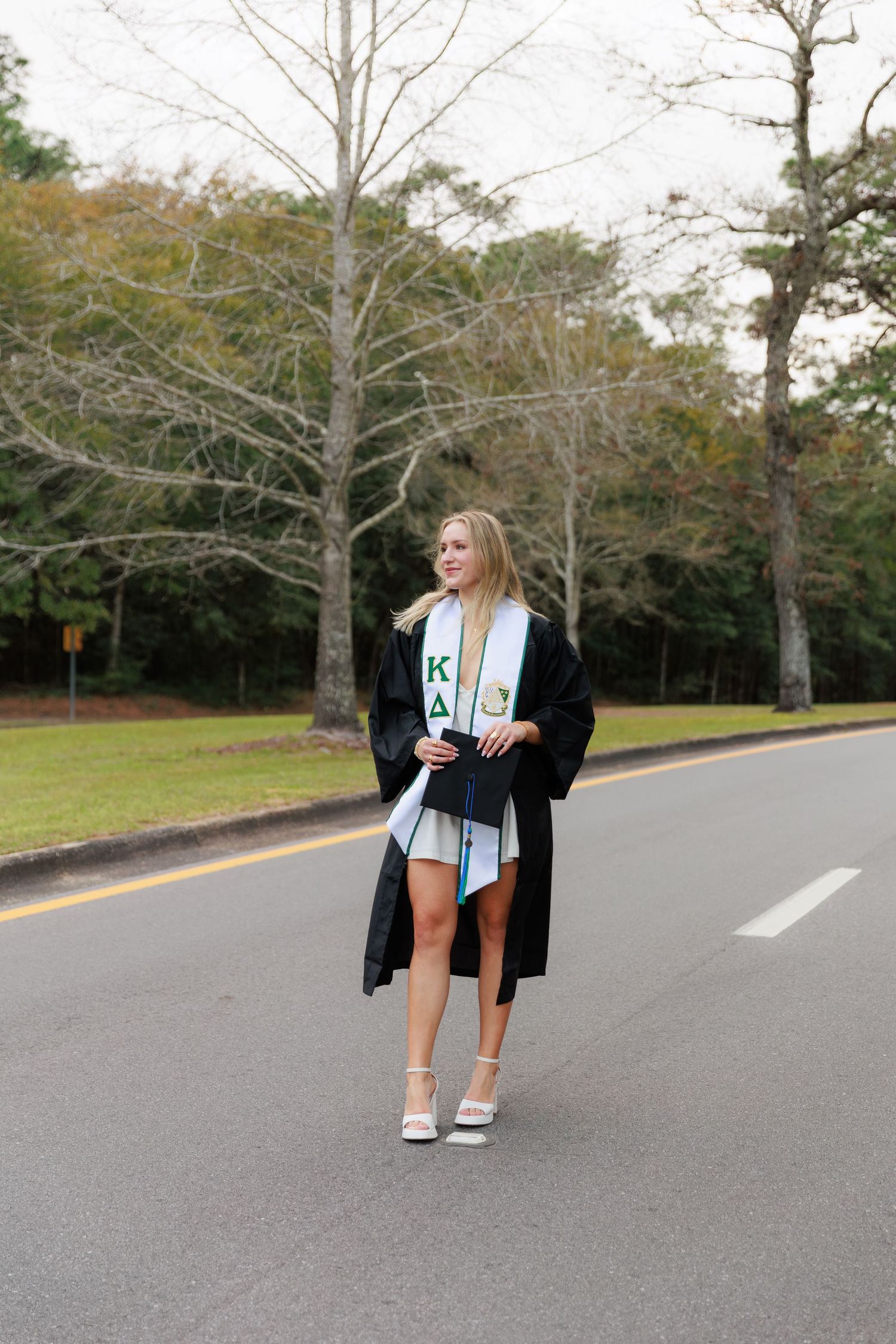 Female graduate in black gown and Kappa Delta sorority stole posing on tree-lined campus road.
