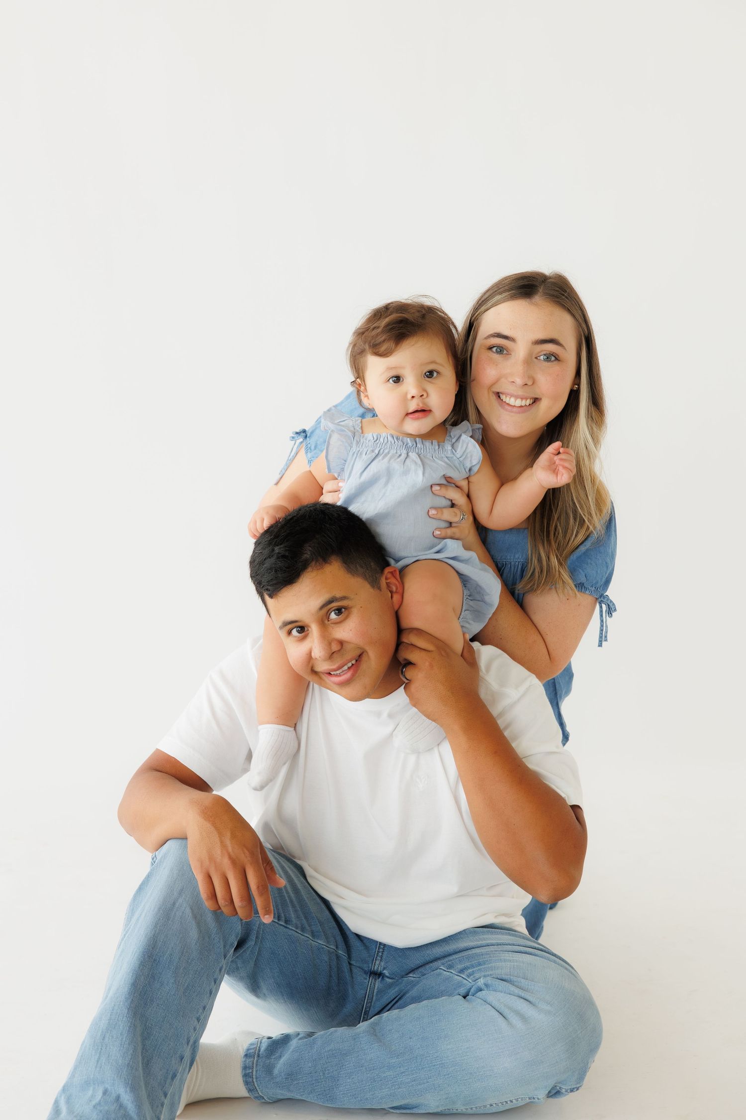 Young family portrait with dad sitting, mom standing behind holding their baby girl, all wearing denim and white outfits.