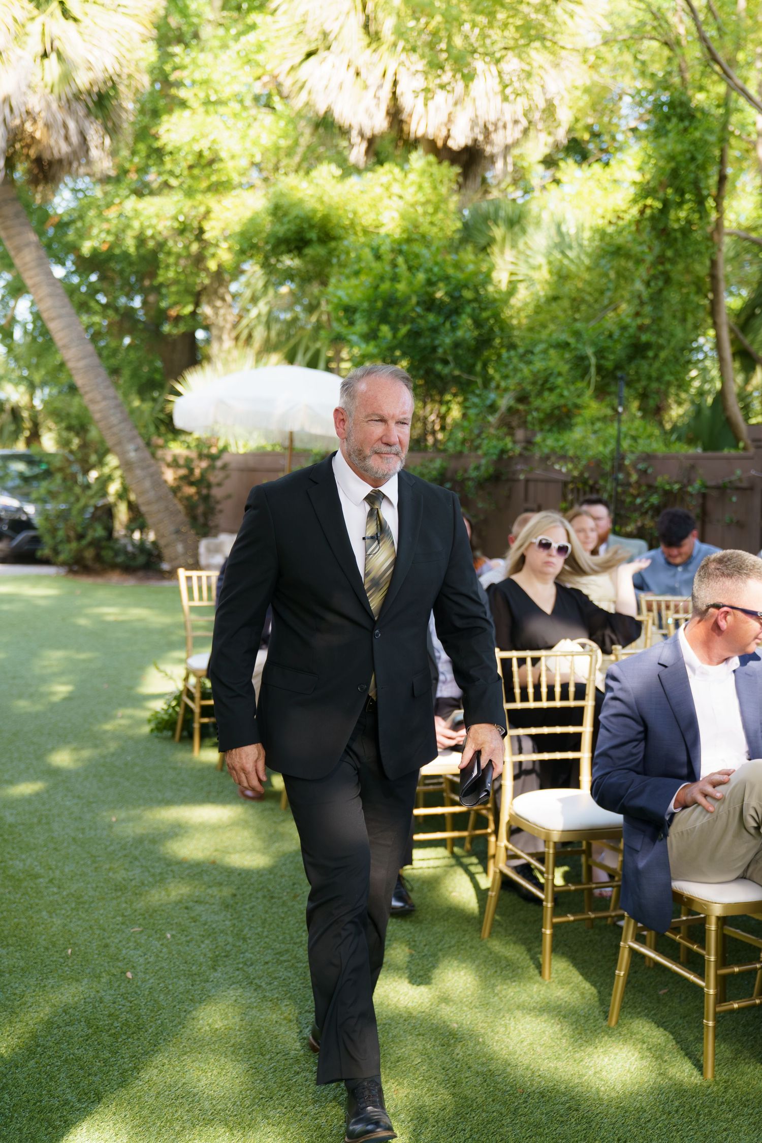 Man in black suit walks down outdoor wedding aisle on green grass.