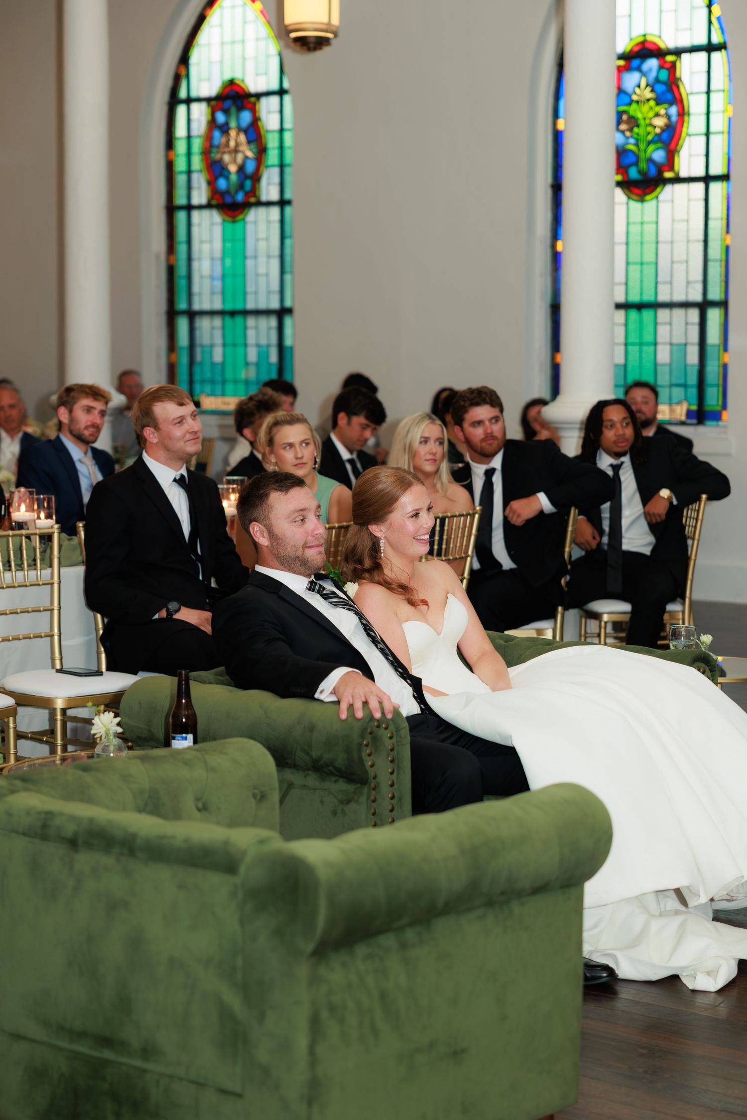 Bride and groom sit together on a green velvet sofa listening to speeches at their elegant chapel wedding reception.