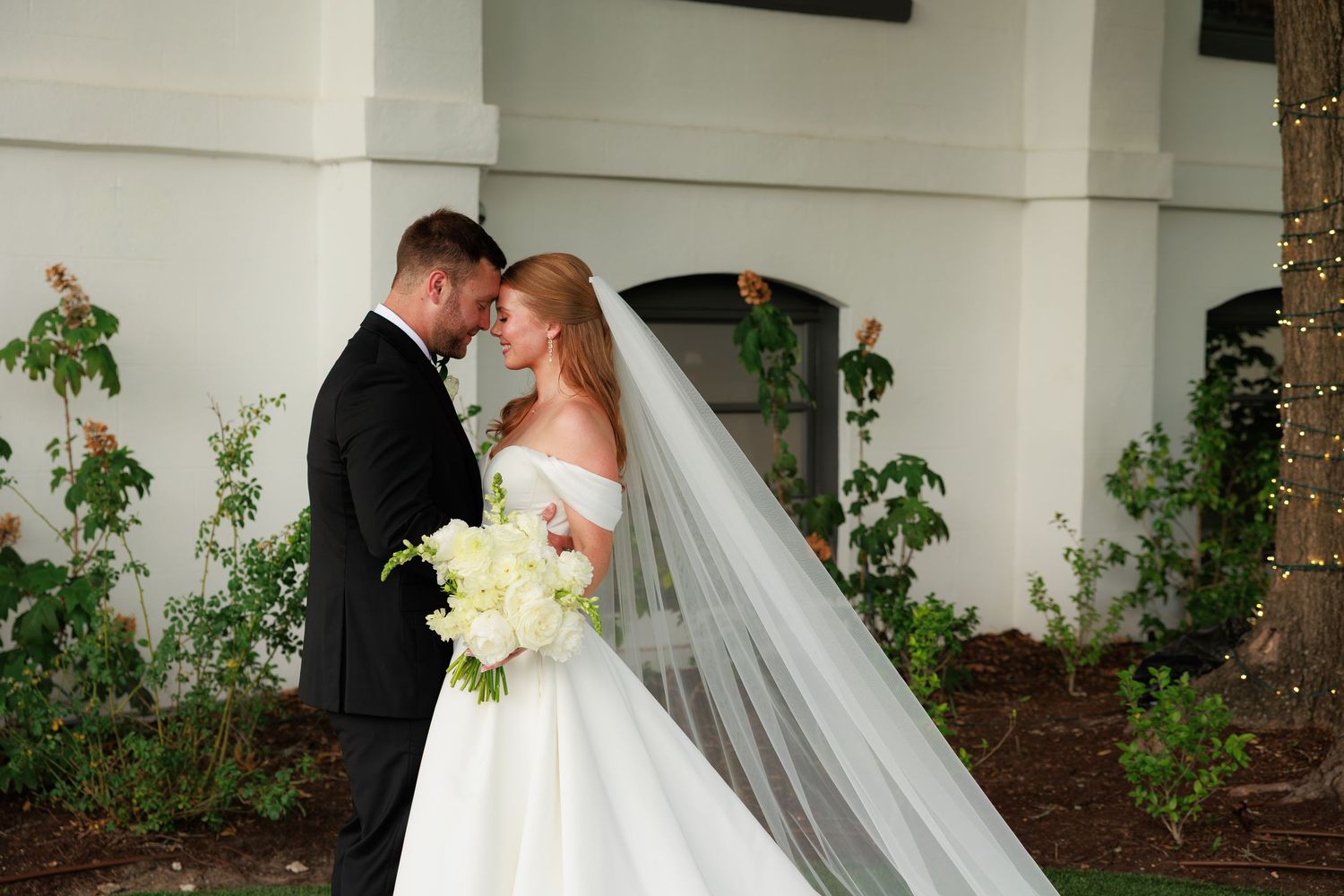 Bride in white ball gown and veil holds bouquet while sharing romantic moment with groom in black tuxedo outdoors.
