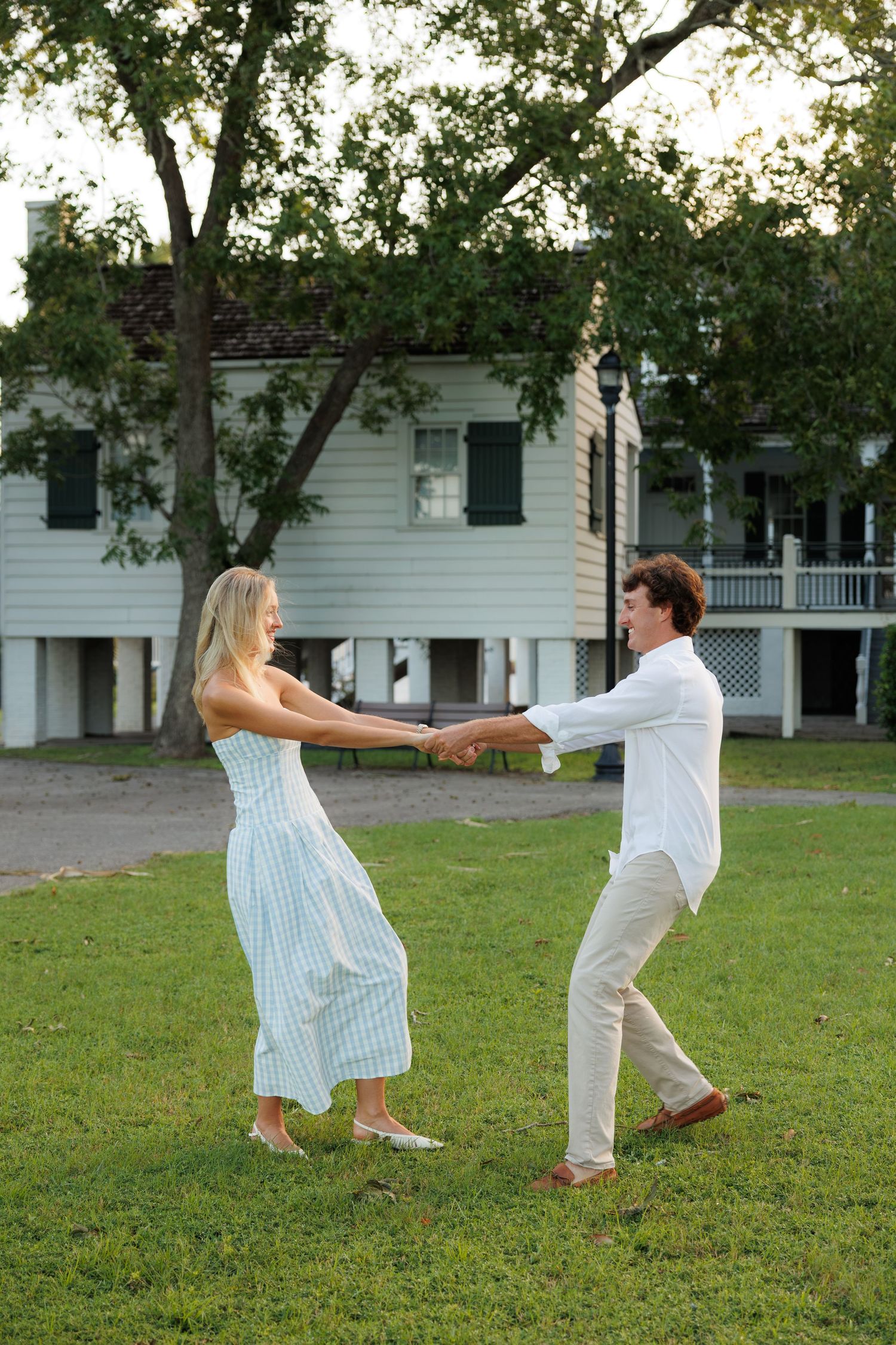 A couple joyfully holds hands and dances on a green lawn in front of a white historic building.