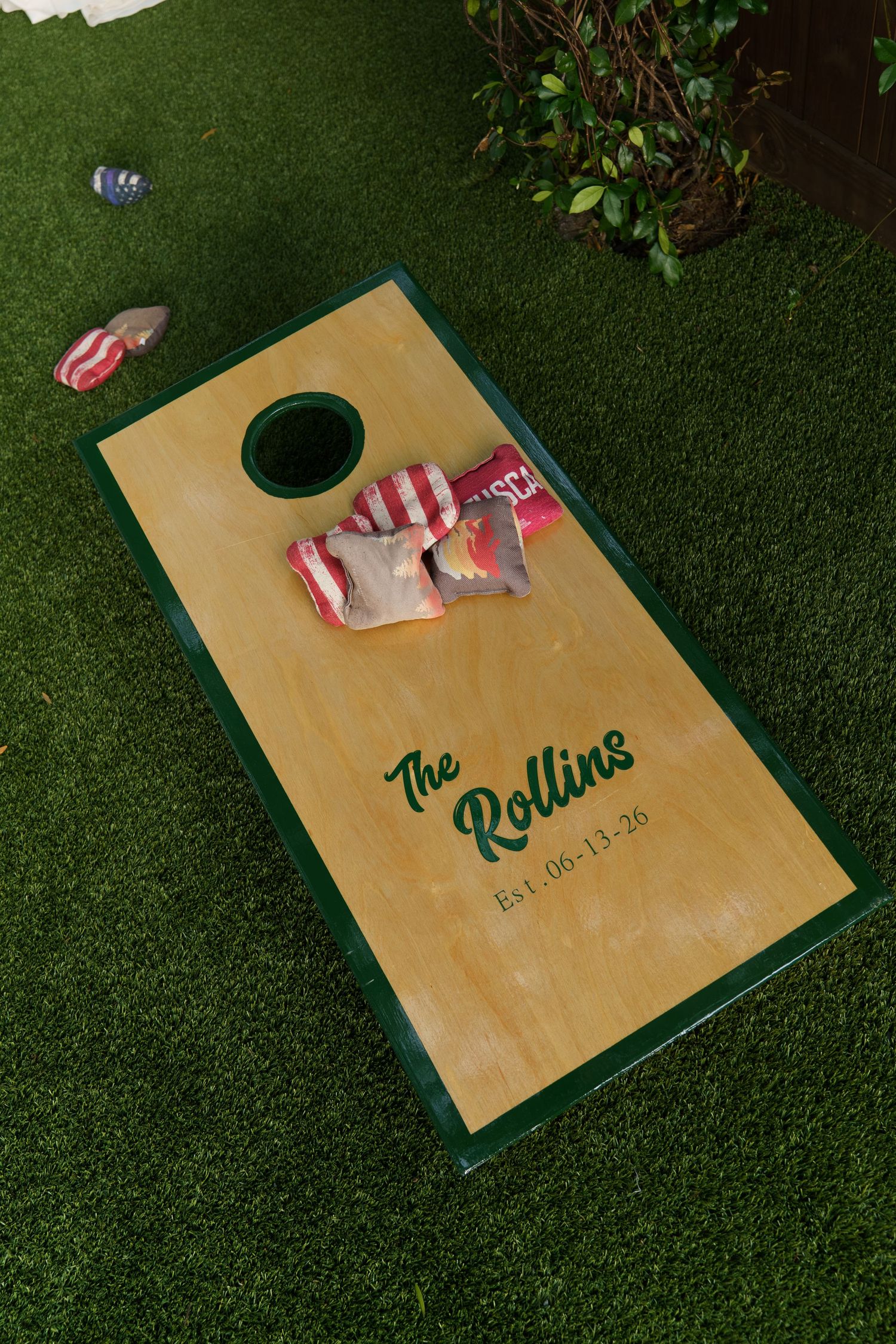 Custom cornhole board reading The Rollins Est. 06-13-26 with red striped bean bags on green artificial grass.
