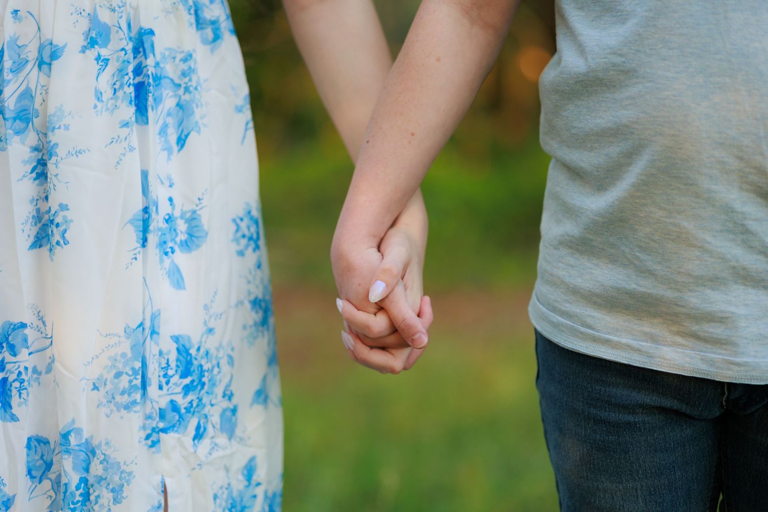 Couple holding hands outdoors, woman in floral dress and man in gray shirt, with green bokeh background.