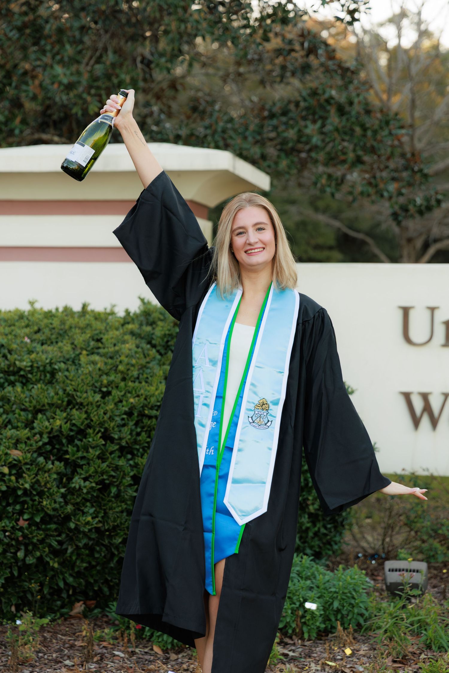 Graduate in black cap and gown with blue and green graduation stole poses for celebratory photos outdoors.