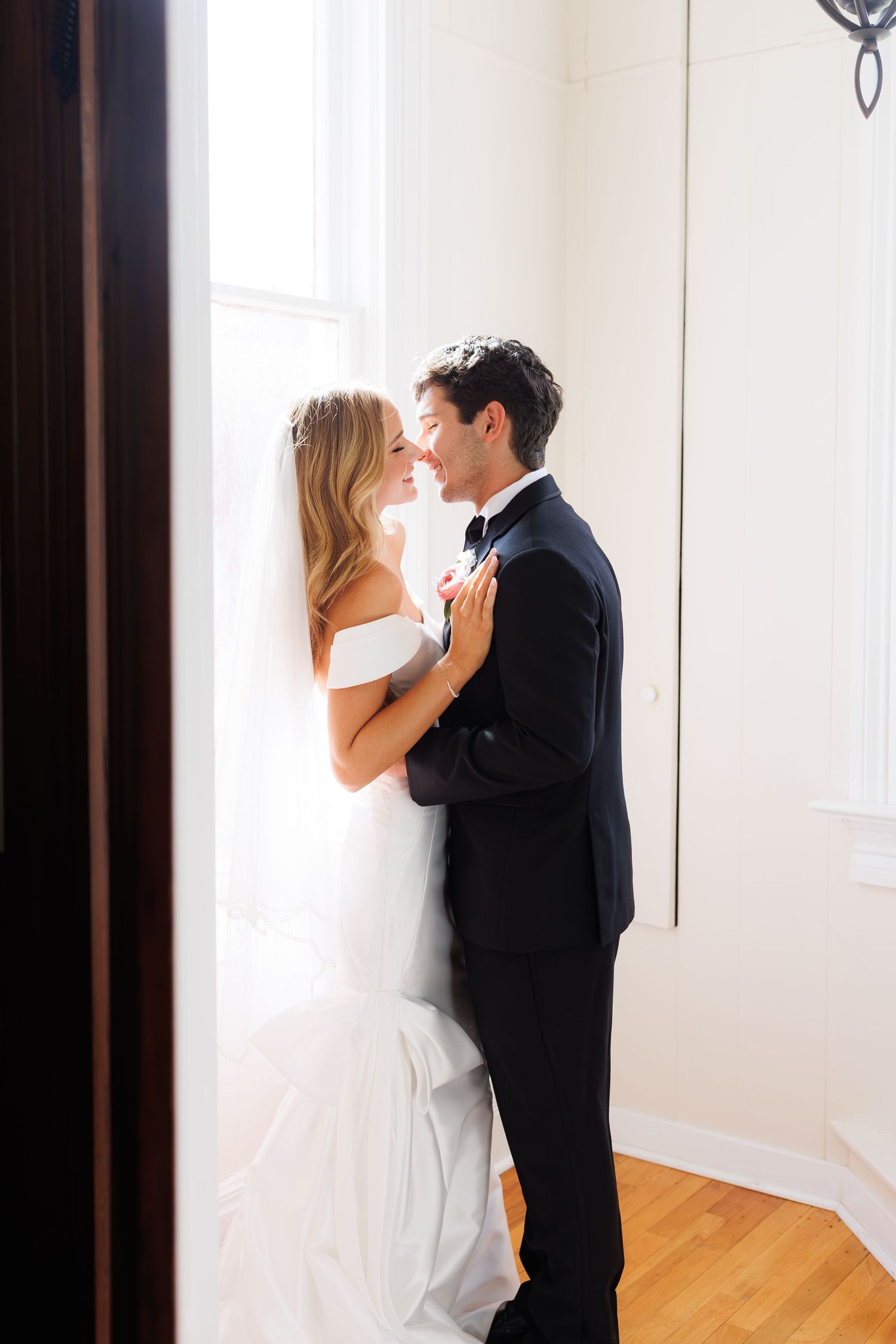A romantic wedding couple shares an intimate moment by a bright window in formal attire.