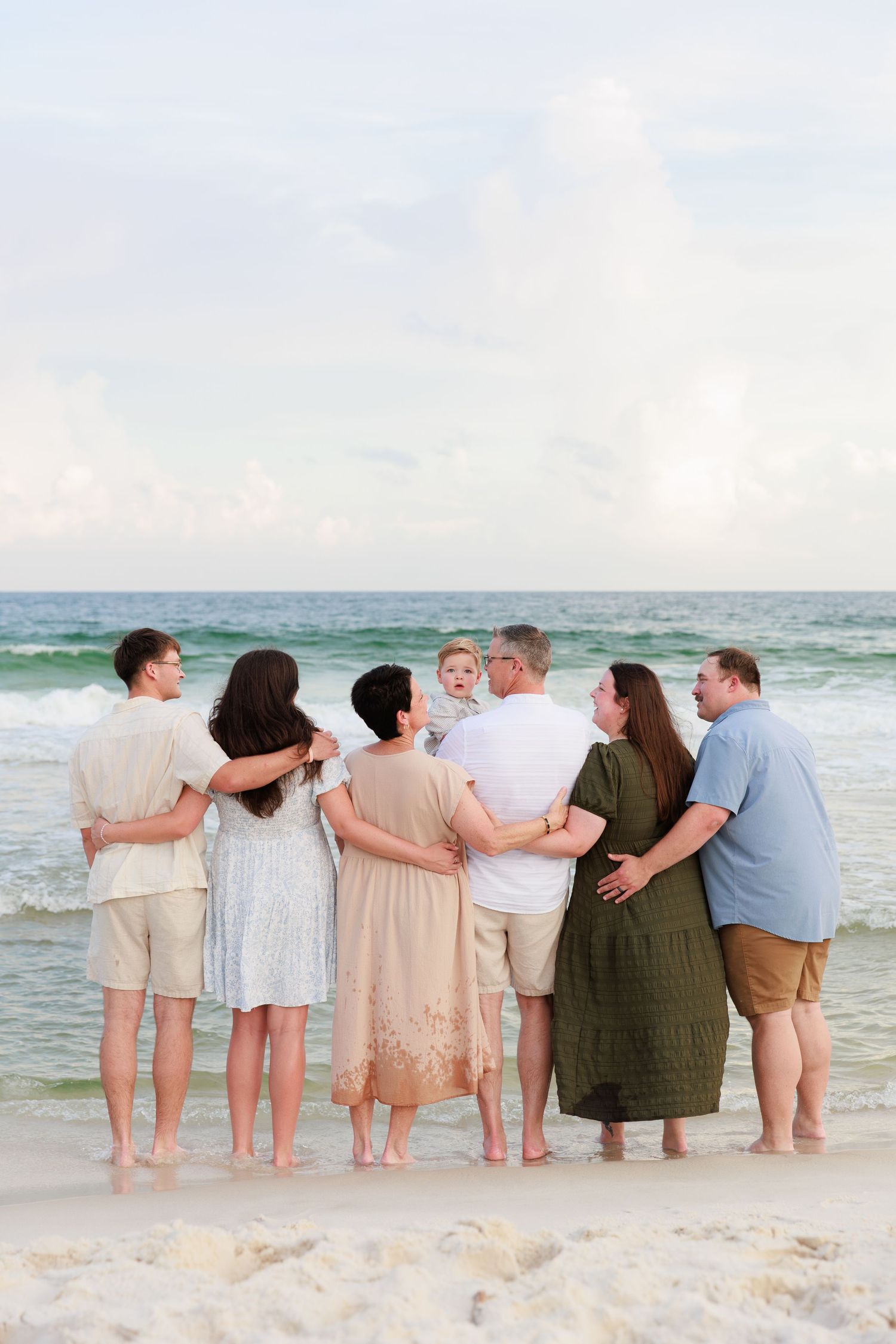 Group of people standing together with arms around each other on a beach at sunset looking at the ocean.