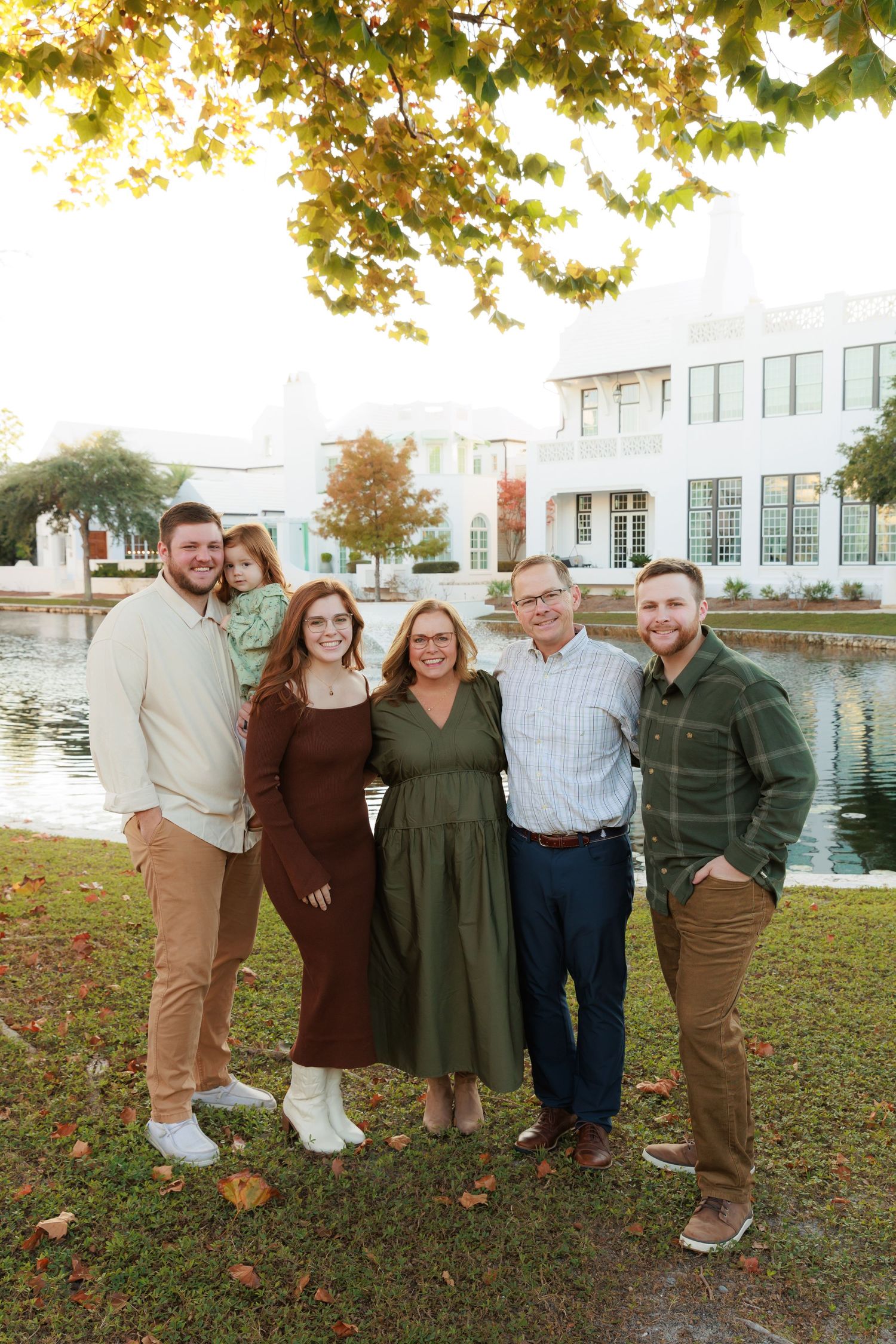 Family group poses together on autumn grass near waterfront with fall foliage and buildings in background.