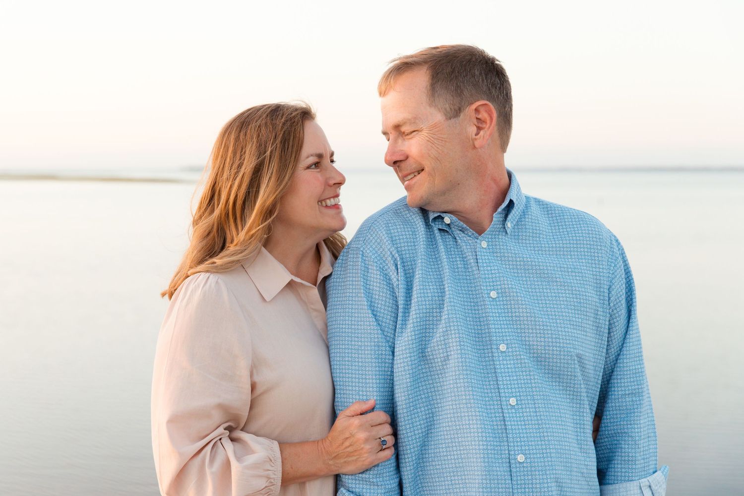 Couple shares intimate moment while standing near calm waterfront during sunset in casual attire.