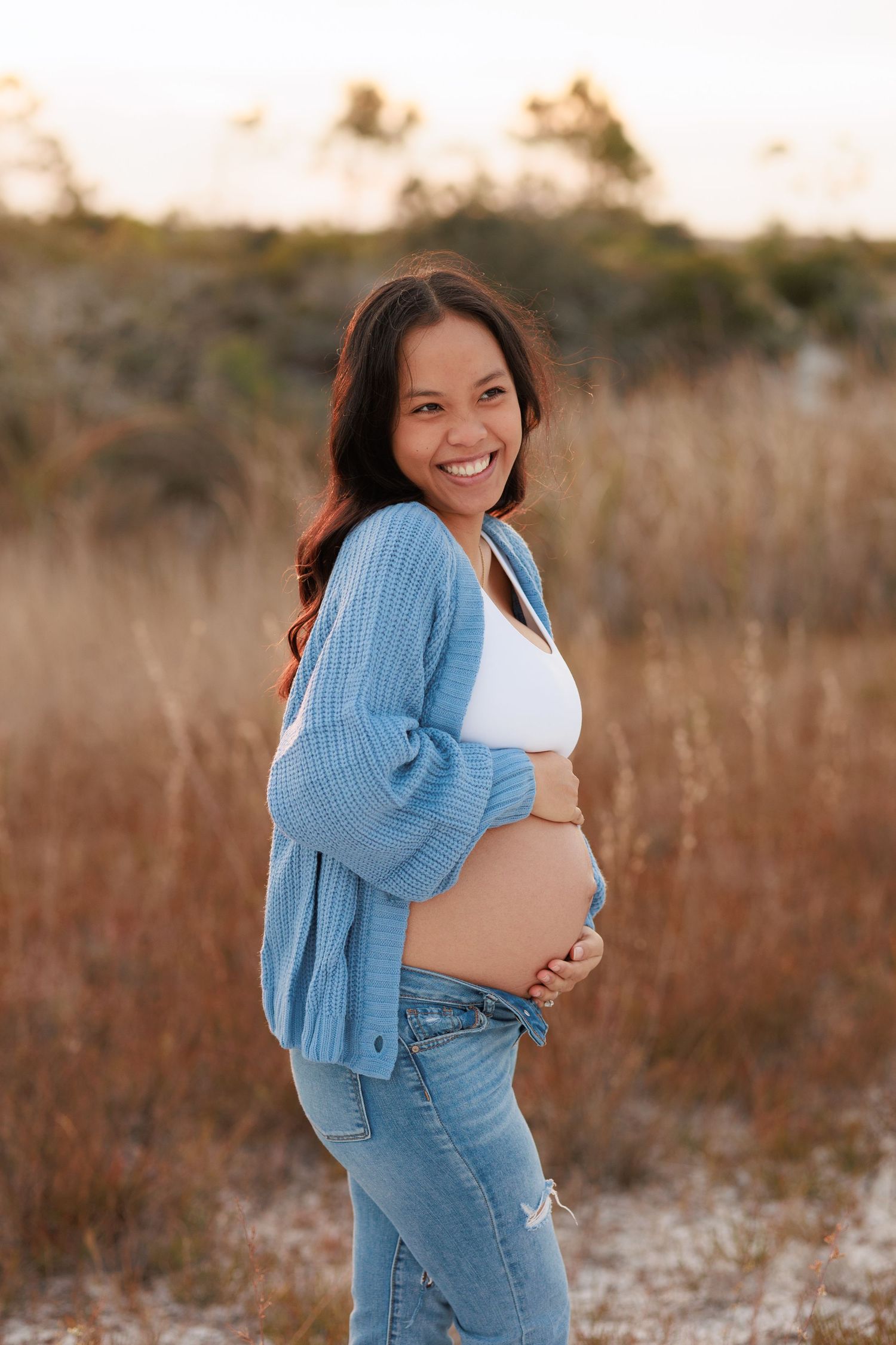 Maternity photo shoot in a field showing a pregnant woman wearing a blue cardigan and white top at sunset.