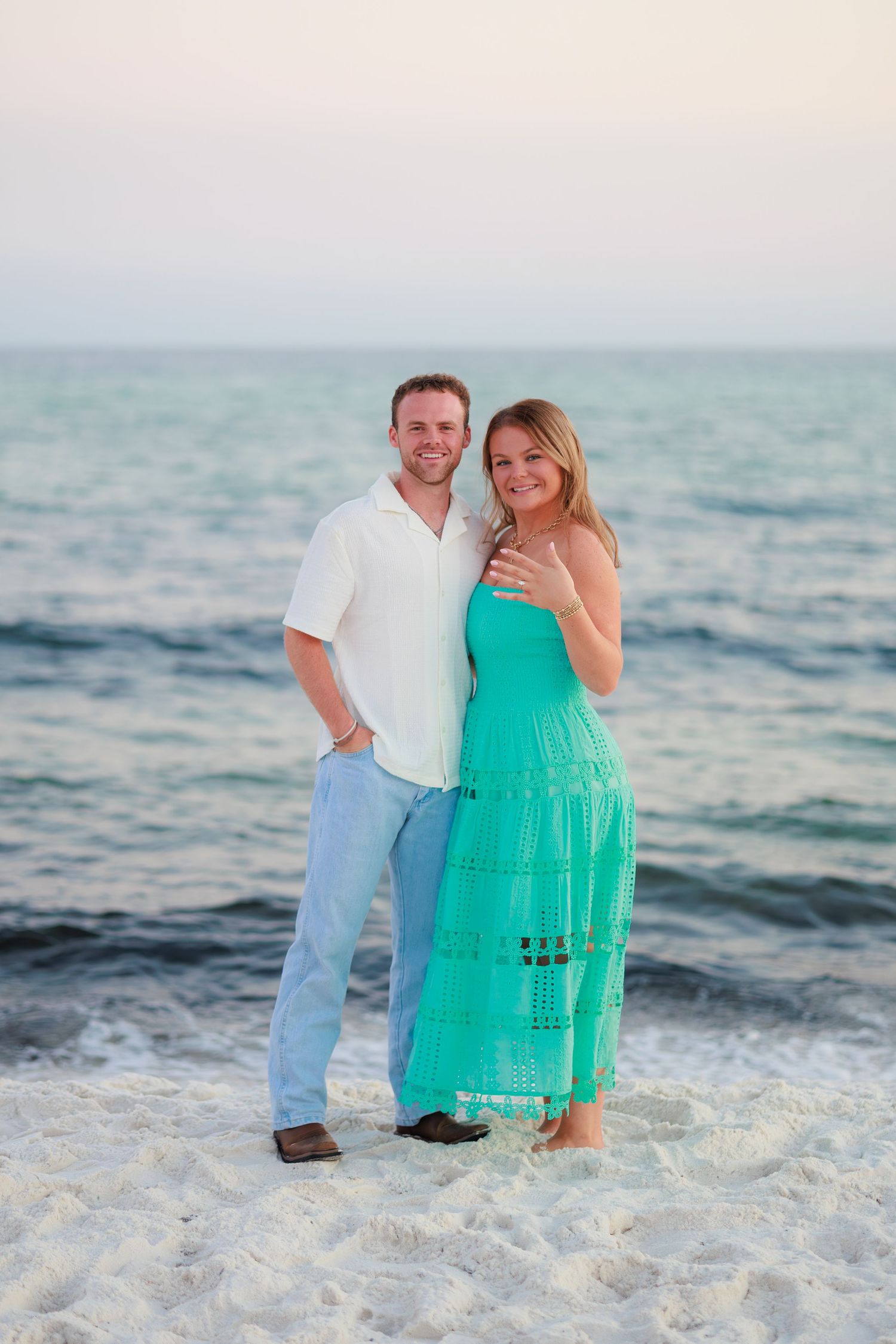 A couple in beachwear poses together on white sand with ocean waves in the background during sunset.
