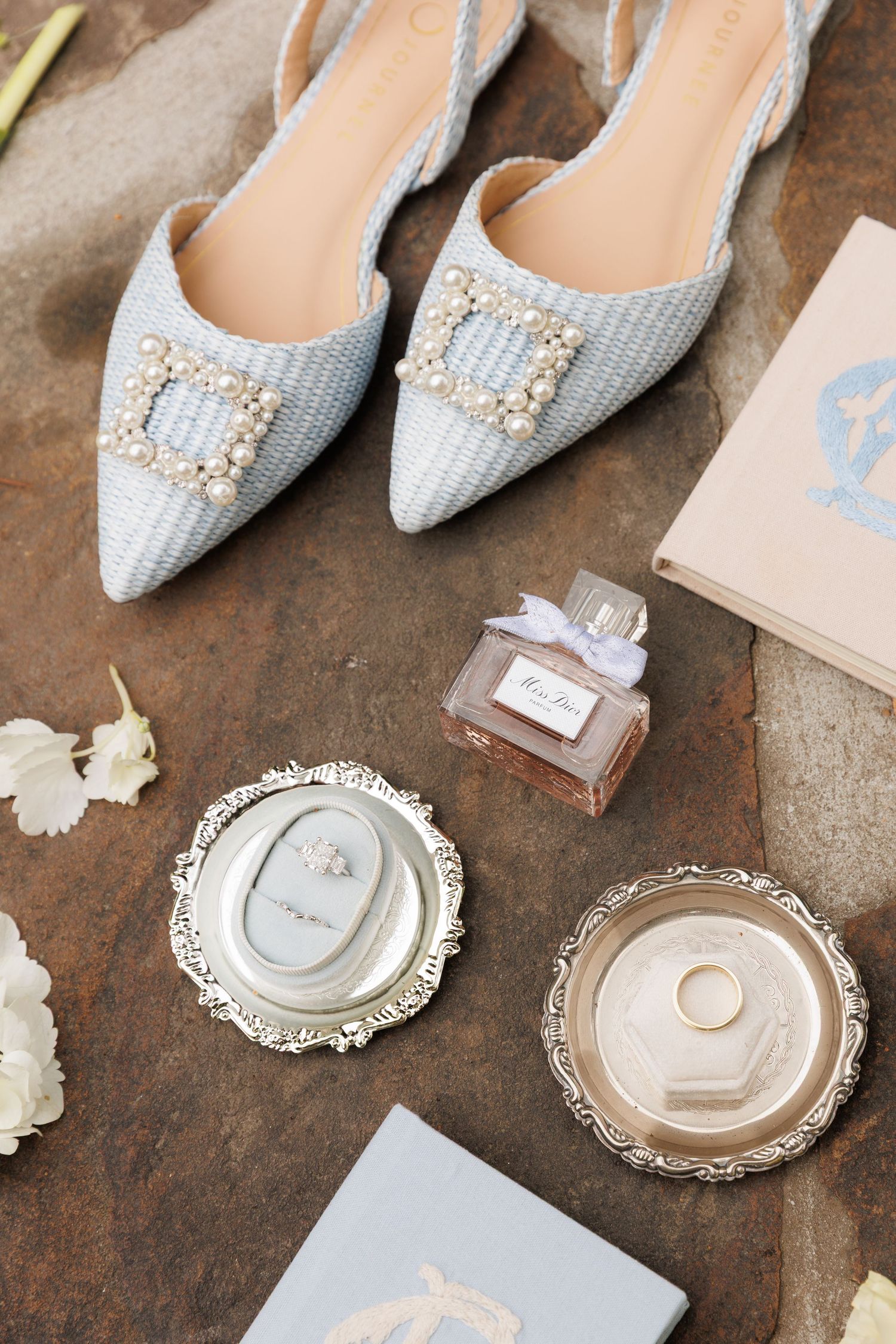 Elegant bridal flat shoes with pearl buckles beside Miss Dior perfume and silver ring dishes on stone.