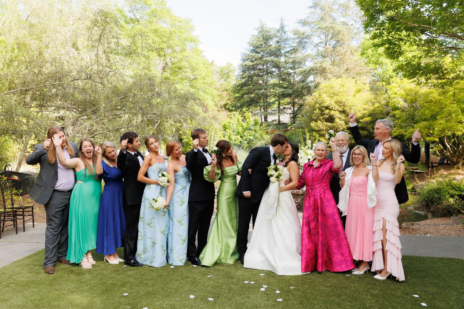 Large wedding party celebrating outdoors with bridesmaids in colorful gowns as bride and groom share a kiss.