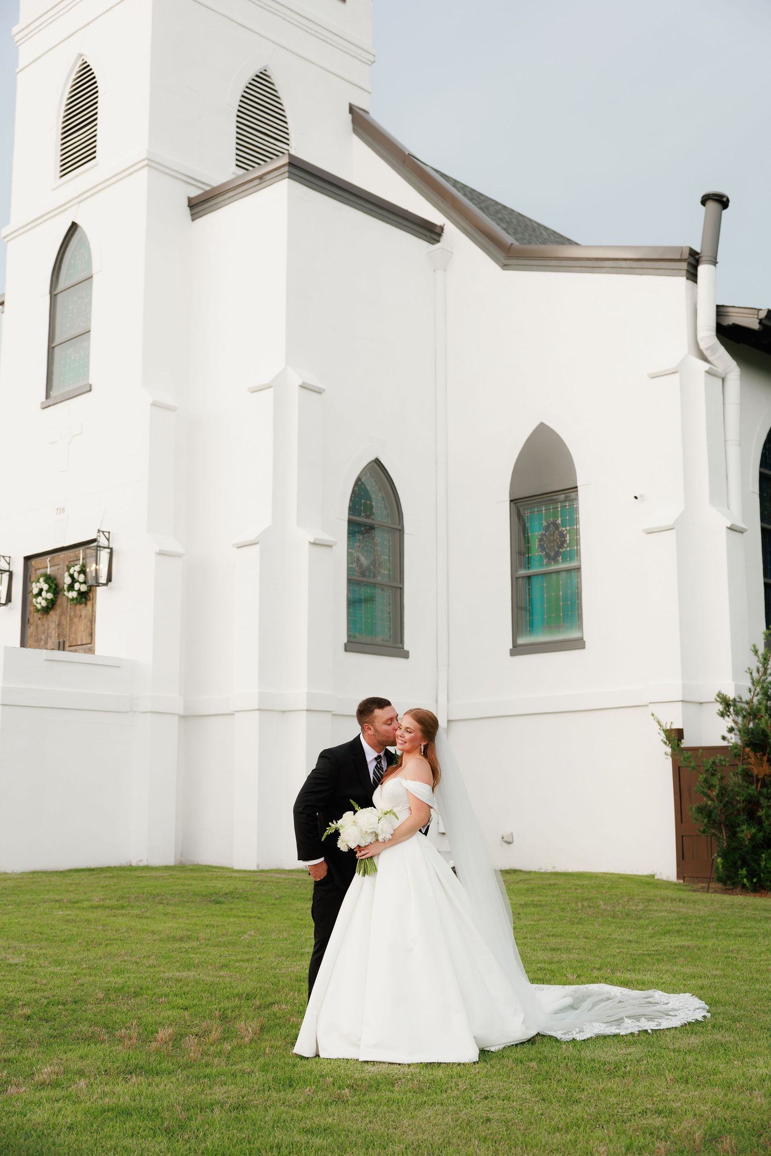 Bride and groom kissing beside white church, bride in flowing off-shoulder gown with cathedral veil on green lawn.