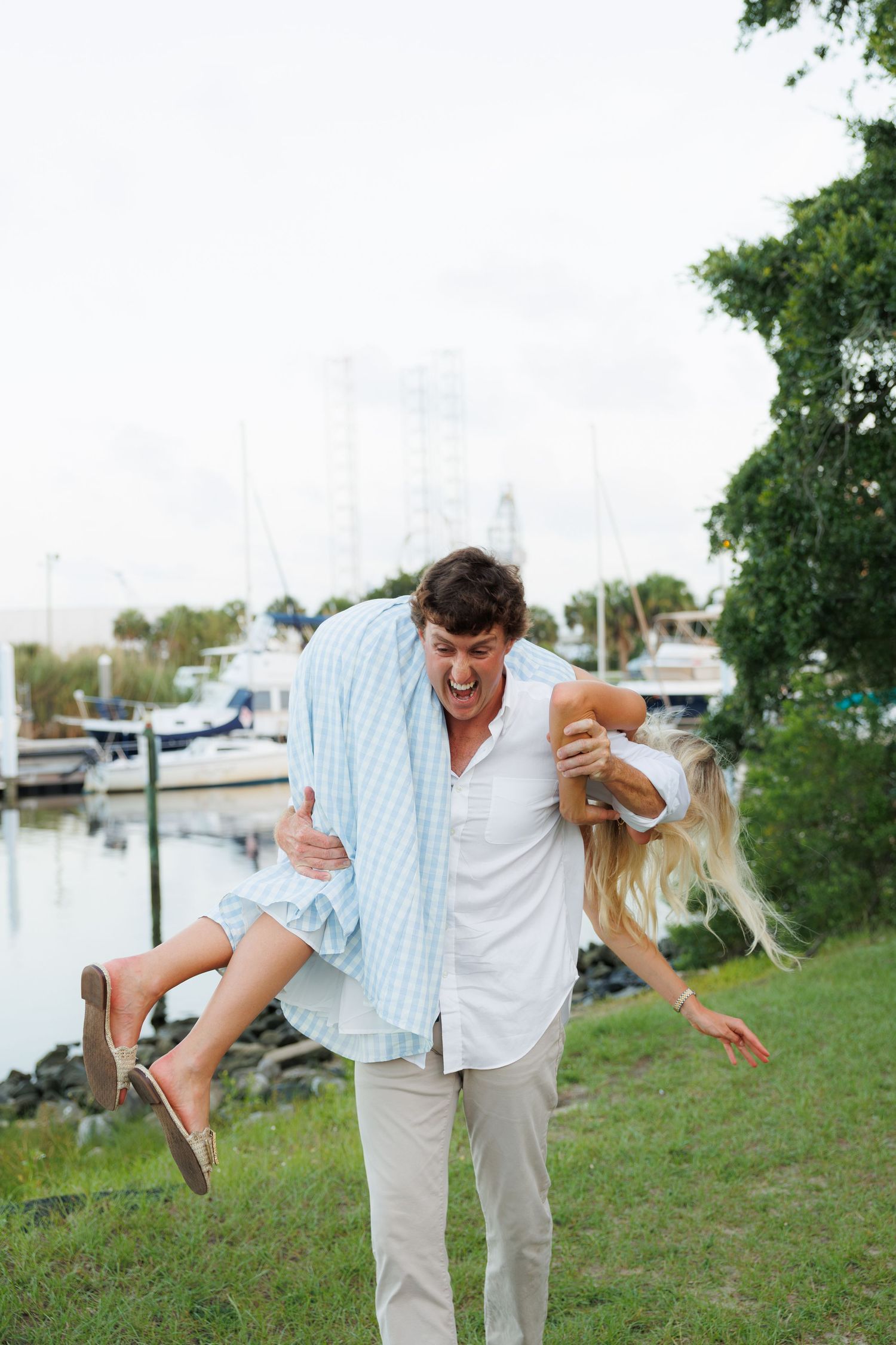 A laughing man carries a woman over his shoulder near a marina with boats and lush greenery in the background.
