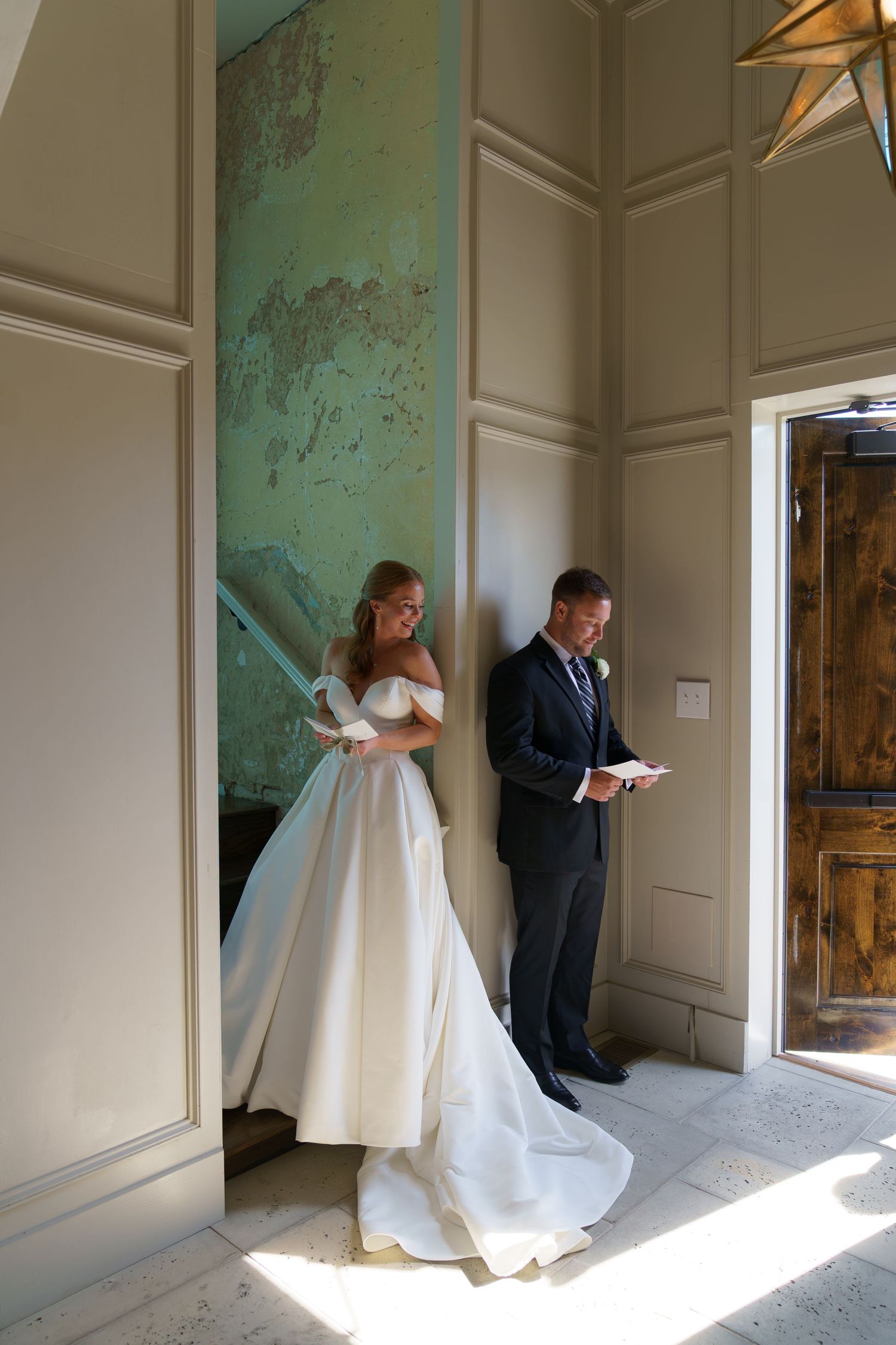 Bride in off-shoulder gown and groom in navy suit read wedding letters back-to-back in elegant venue.