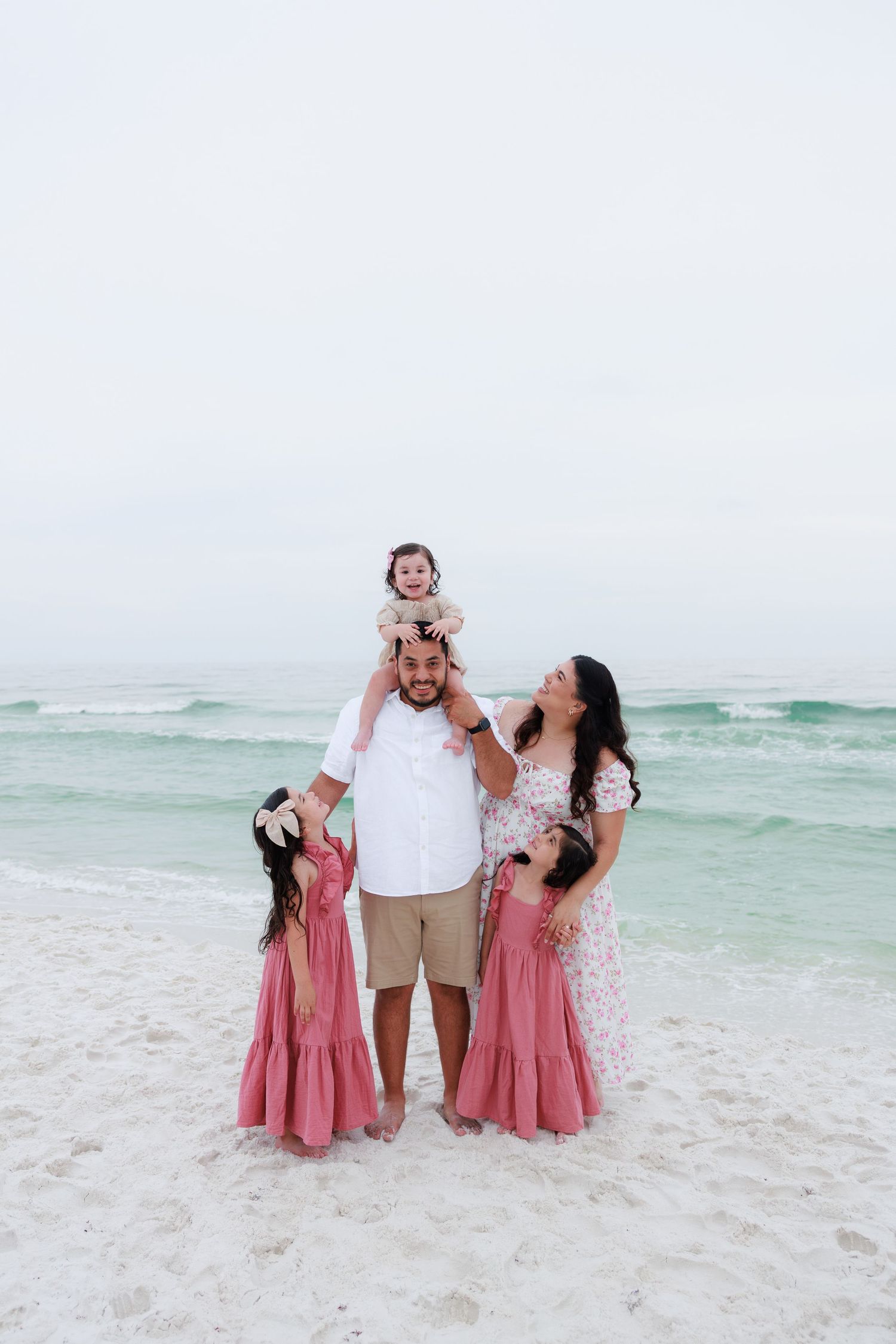 Family of five posing on white sand beach, parents with three young daughters in matching pink maxi dresses, ocean backdrop.