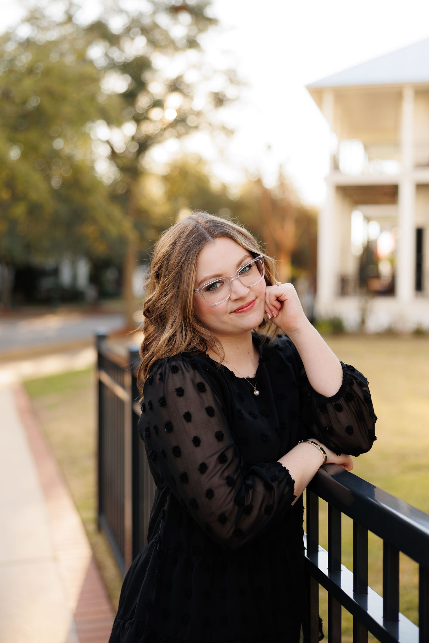 Young woman in black polka dot dress and glasses leaning on railing outdoors near a white building.