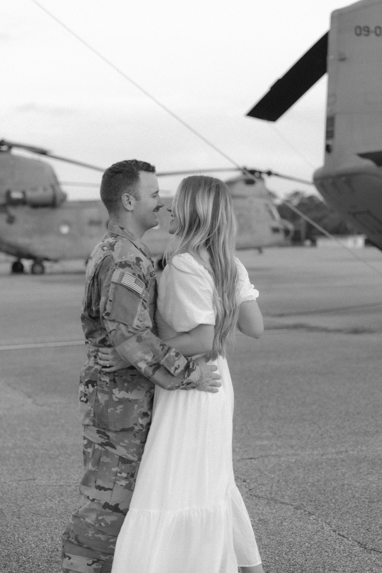 Military couple shares intimate moment in black and white photo near helicopter.