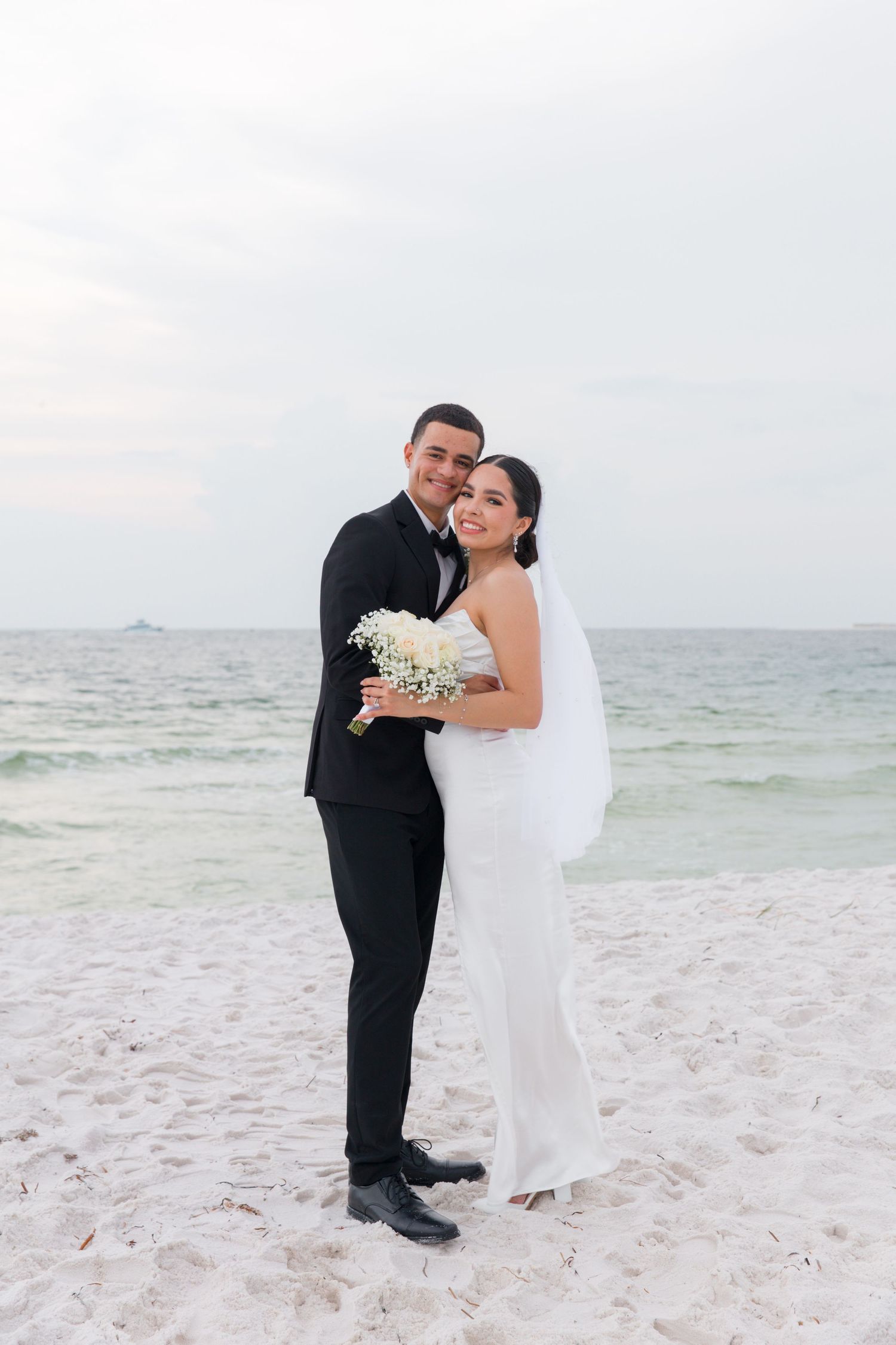 Happy couple poses for wedding photos on pristine beach holding white bouquet.