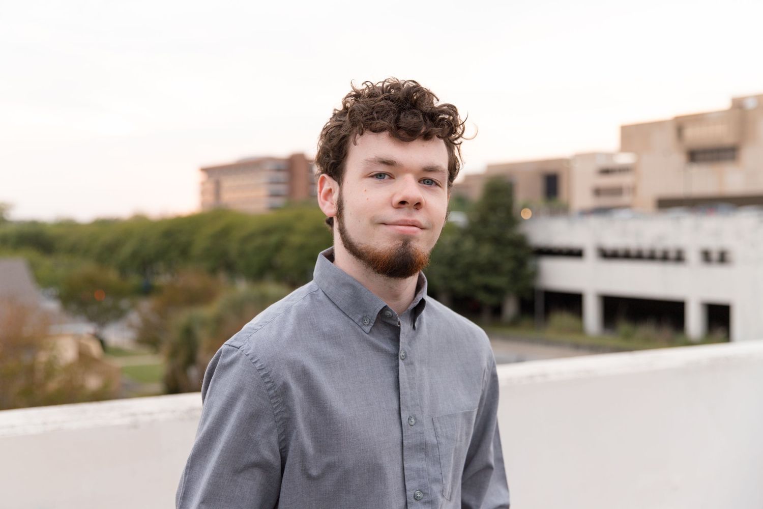 A portrait of a person in a grey button-up shirt standing outside with buildings in the background.