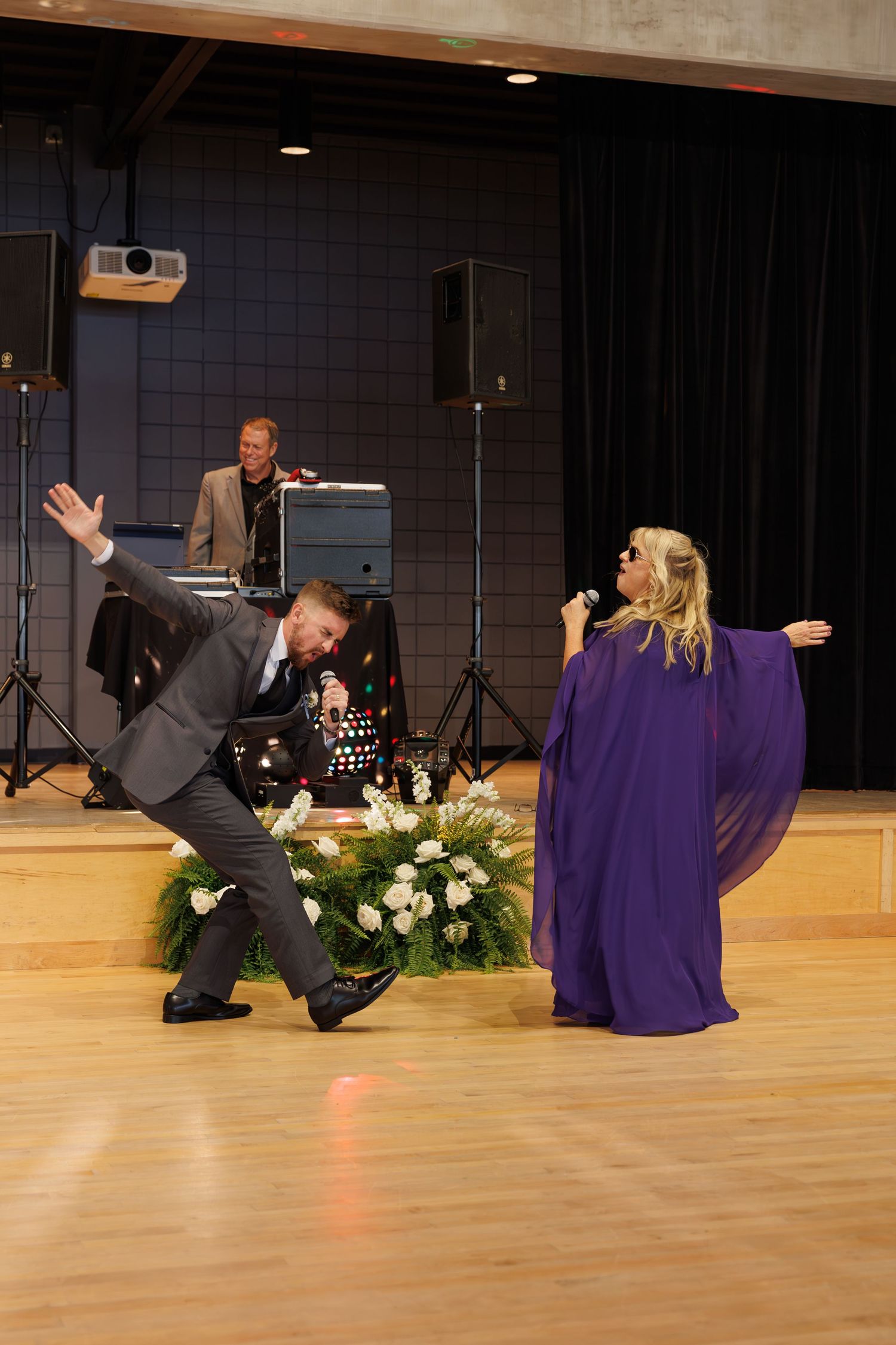 Wedding performers dancing energetically on stage with woman in purple gown and man in grey suit.