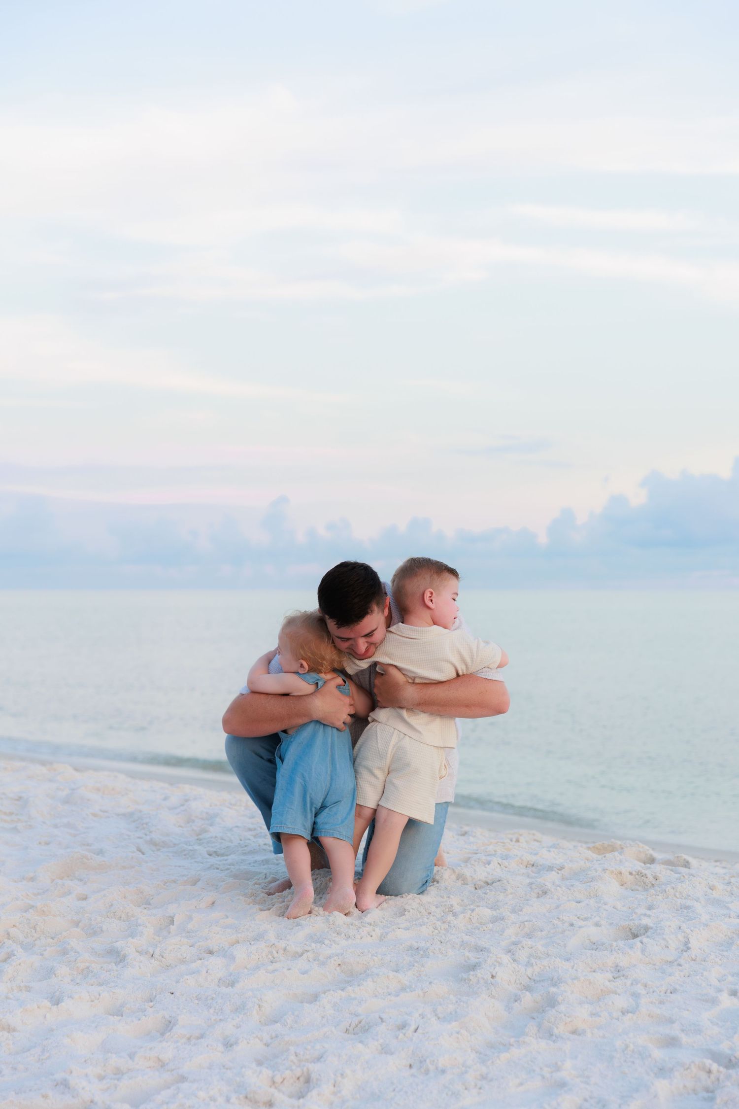 Father kneeling on white sand beach hugging two young children at sunset with calm ocean in background.