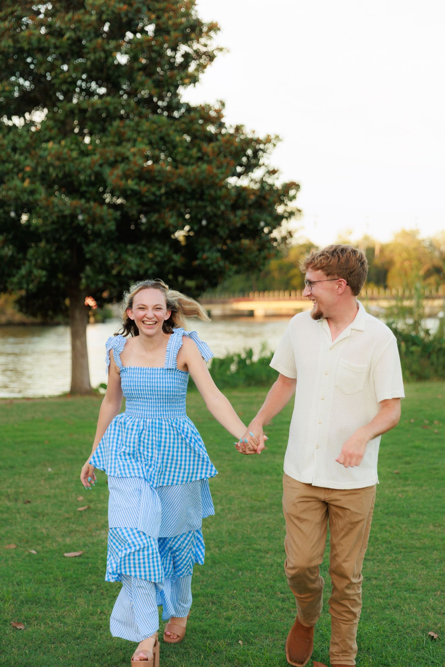 A joyful couple holds hands and runs through green grass near a waterfront, she wears a blue tiered gingham dress.