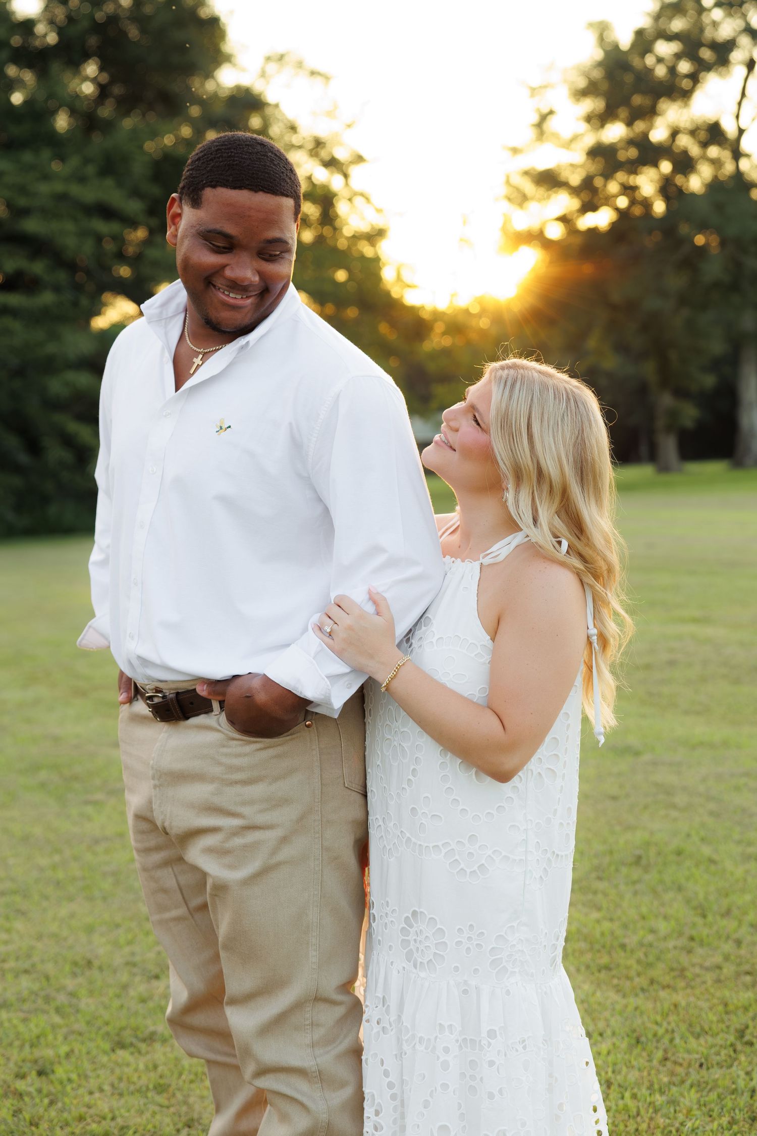 A smiling interracial couple poses at golden hour in a park, man in white shirt and woman in white lace dress.