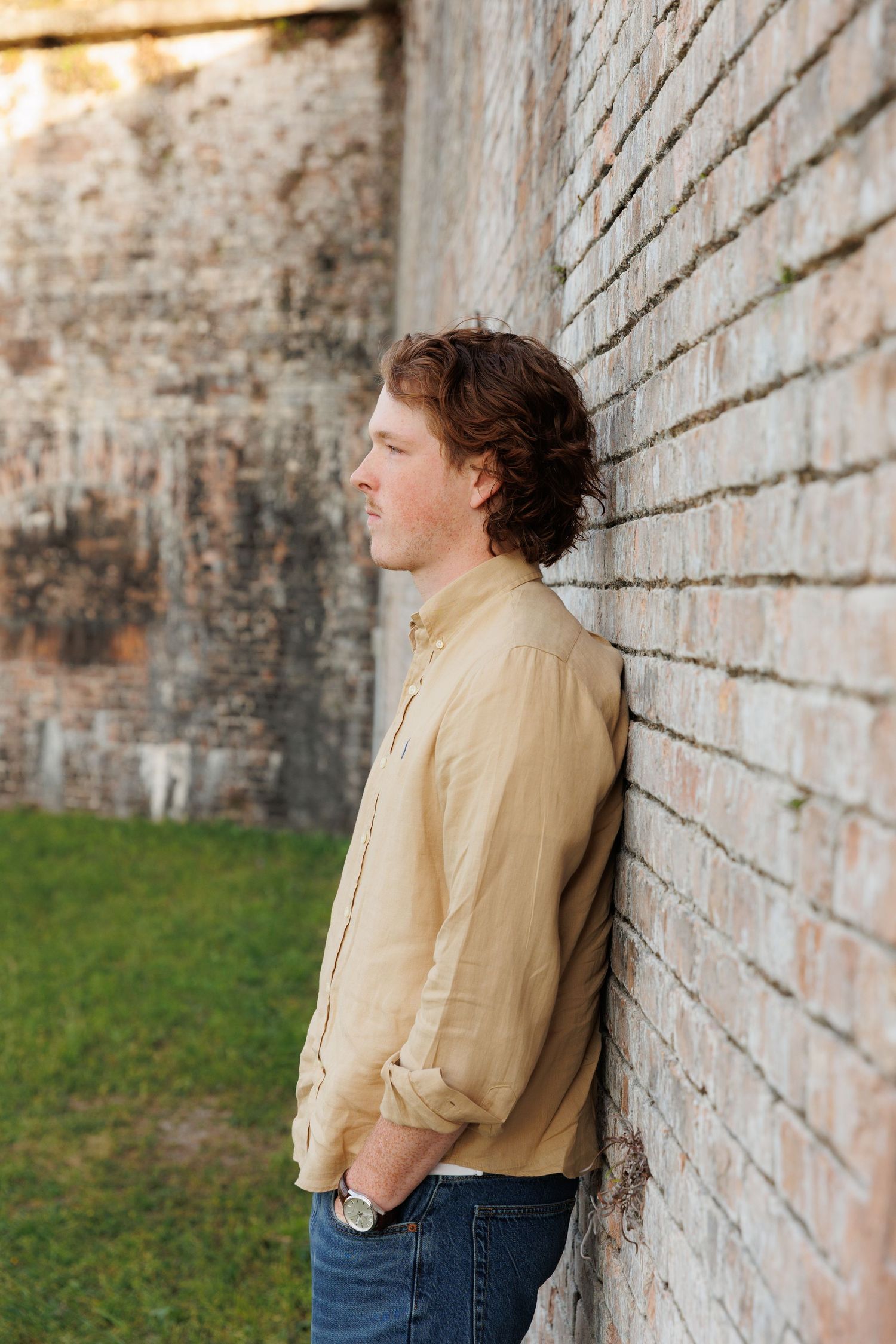 Young man in yellow shirt leaning against brick wall looking sideways outdoors.