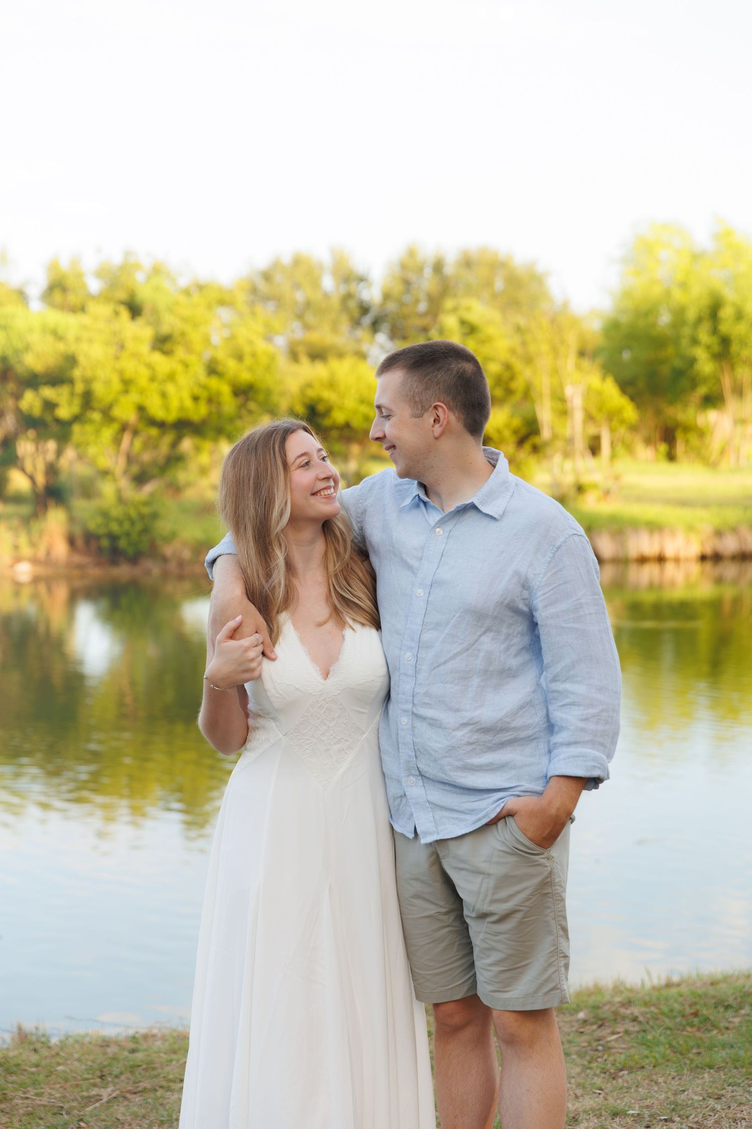 Happy couple laughing together beside a serene pond at sunset, woman in white dress, man in blue shirt.