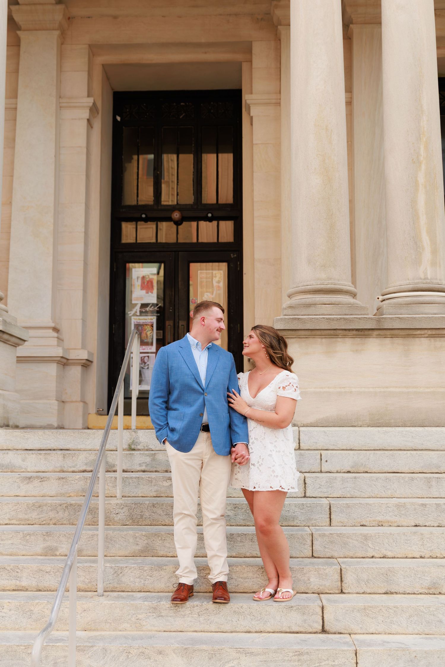 Couple in blue blazer and white lace dress holding hands on stone steps of a classical building.