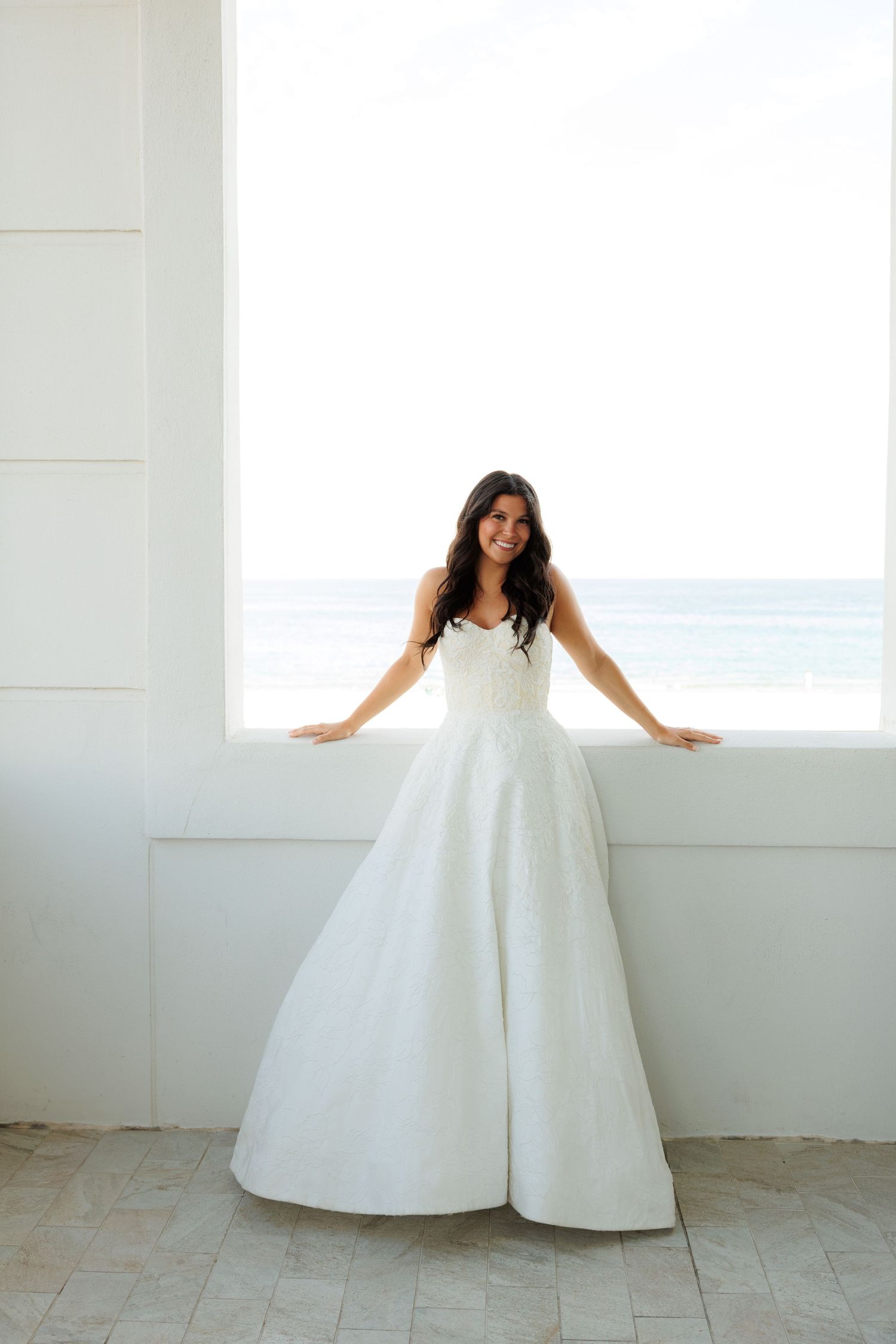Bride in strapless white ball gown wedding dress standing by ocean view window at beachfront venue.