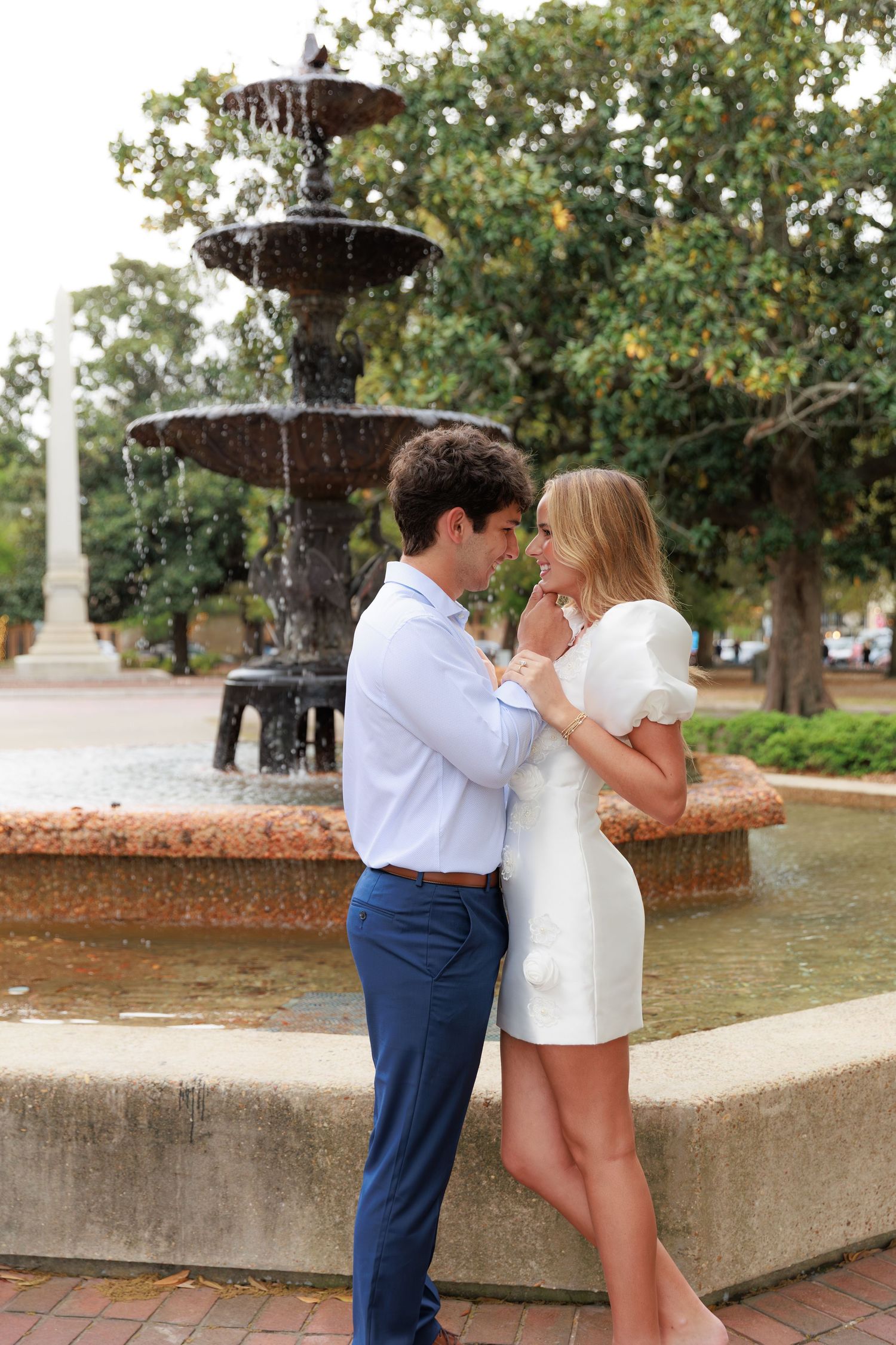 A romantic moment shared between a couple by an ornate tiered fountain in a park setting.