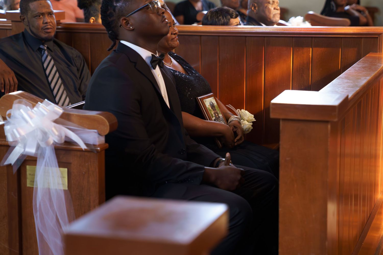 People sit in wooden church pews decorated with white bows during a religious service.